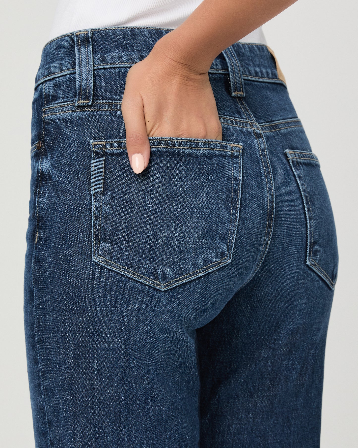 A woman modeling a pair of dark blue high-waisted flare jeans with a white sleeveless top, standing against a light gray studio background.