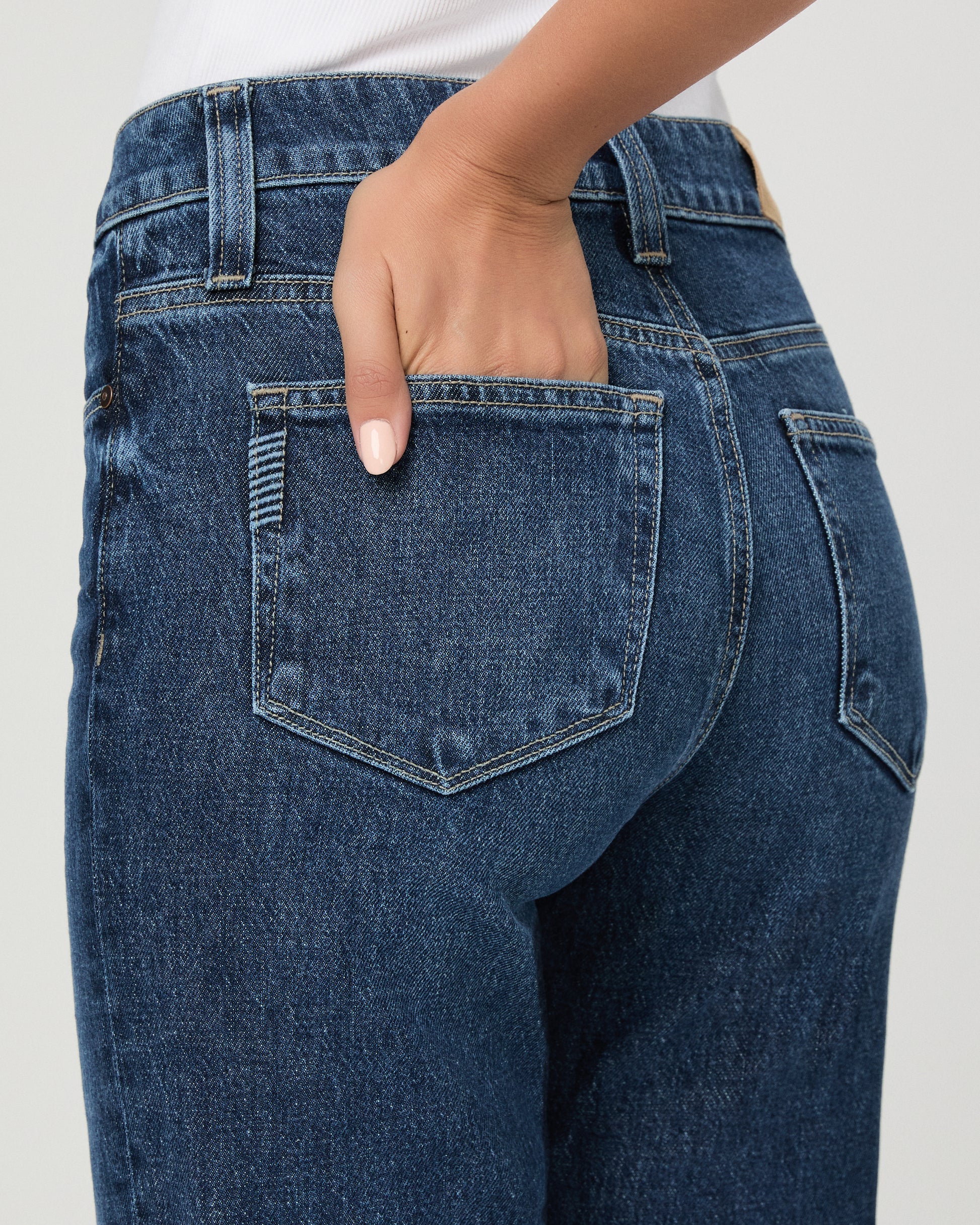 A woman modeling a pair of dark blue high-waisted flare jeans with a white sleeveless top, standing against a light gray studio background.