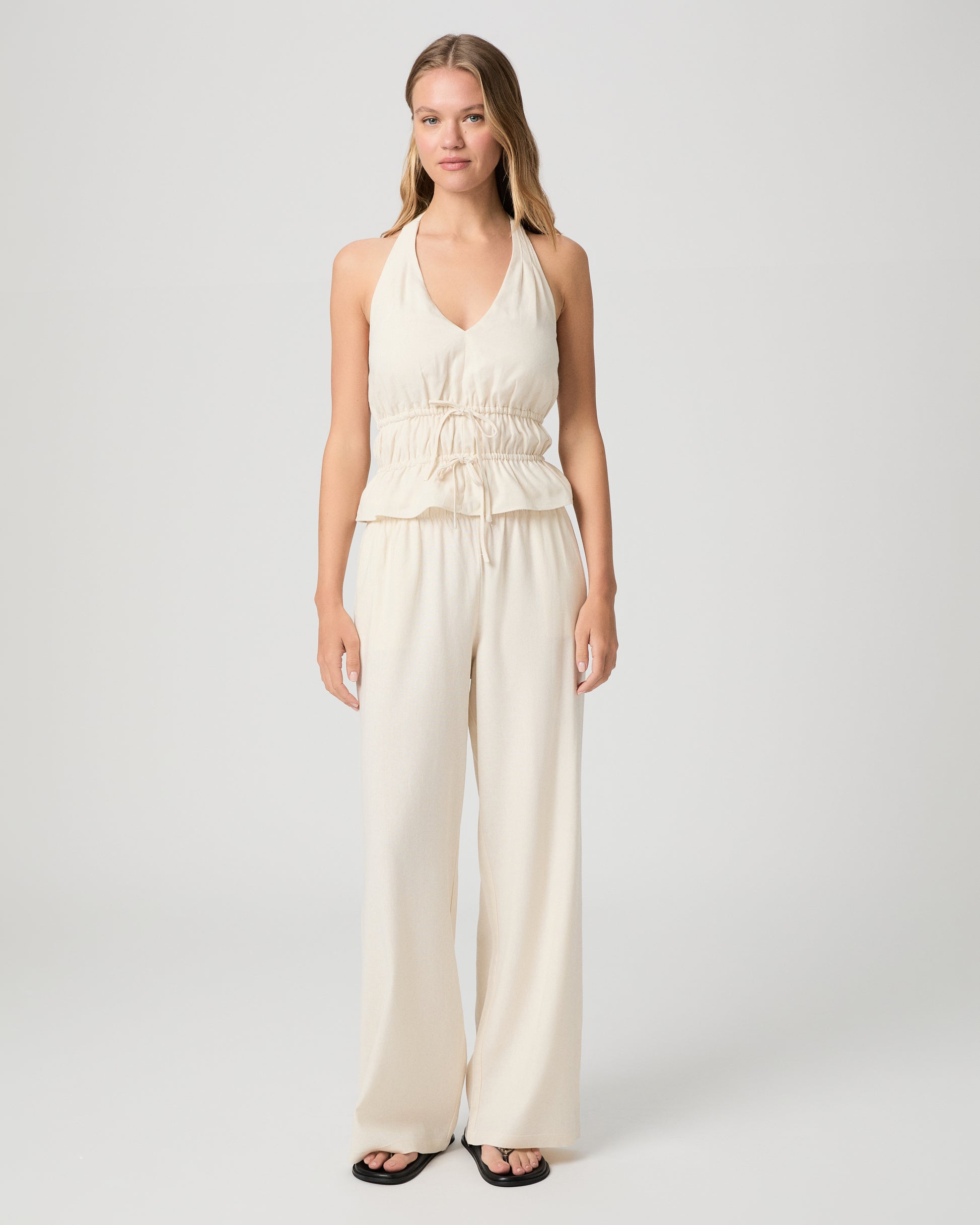 Woman wearing a white halter neck top and white pants against a plain background