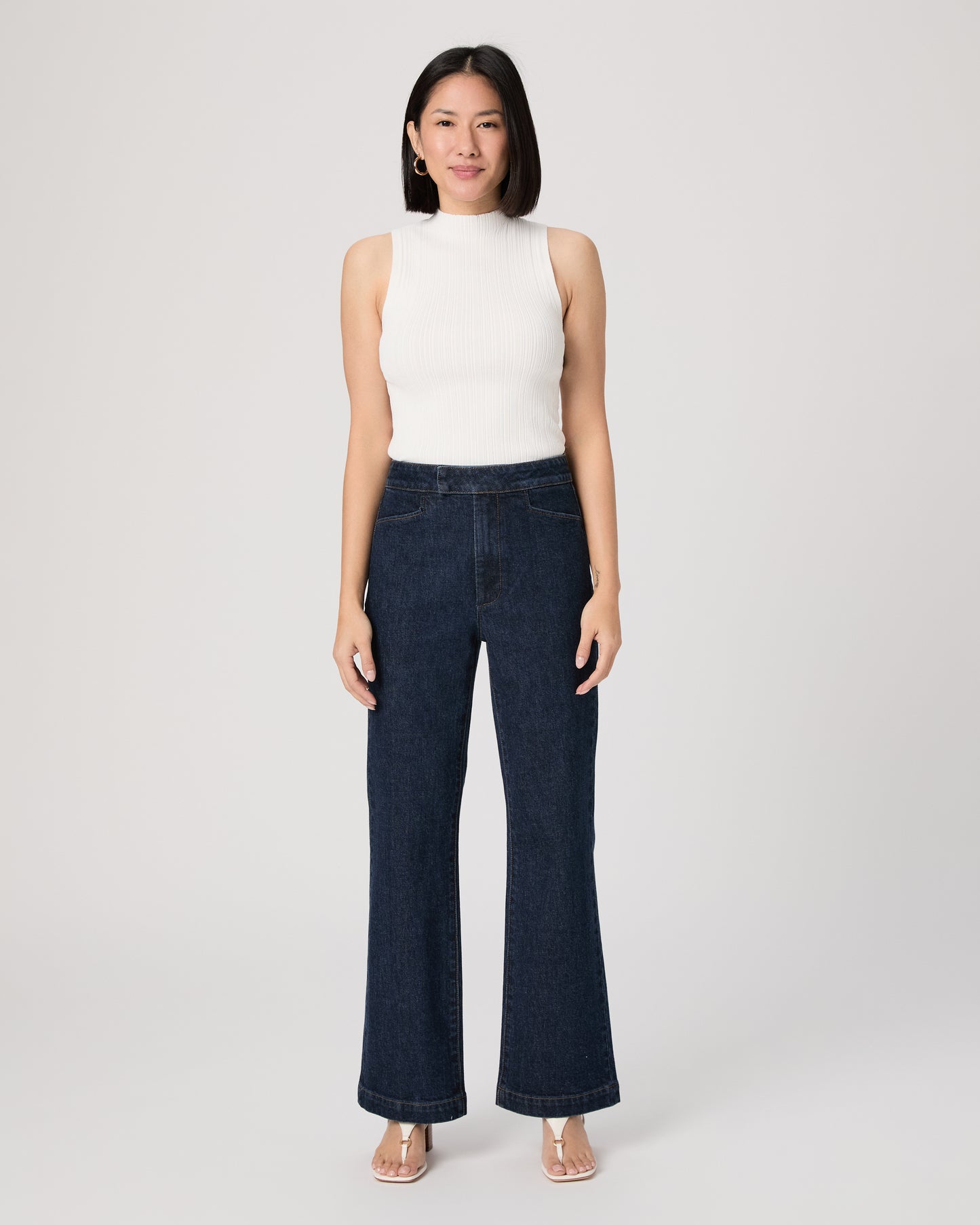 Woman wearing a white sleeveless top and dark blue jeans paired with white sandals on a white background