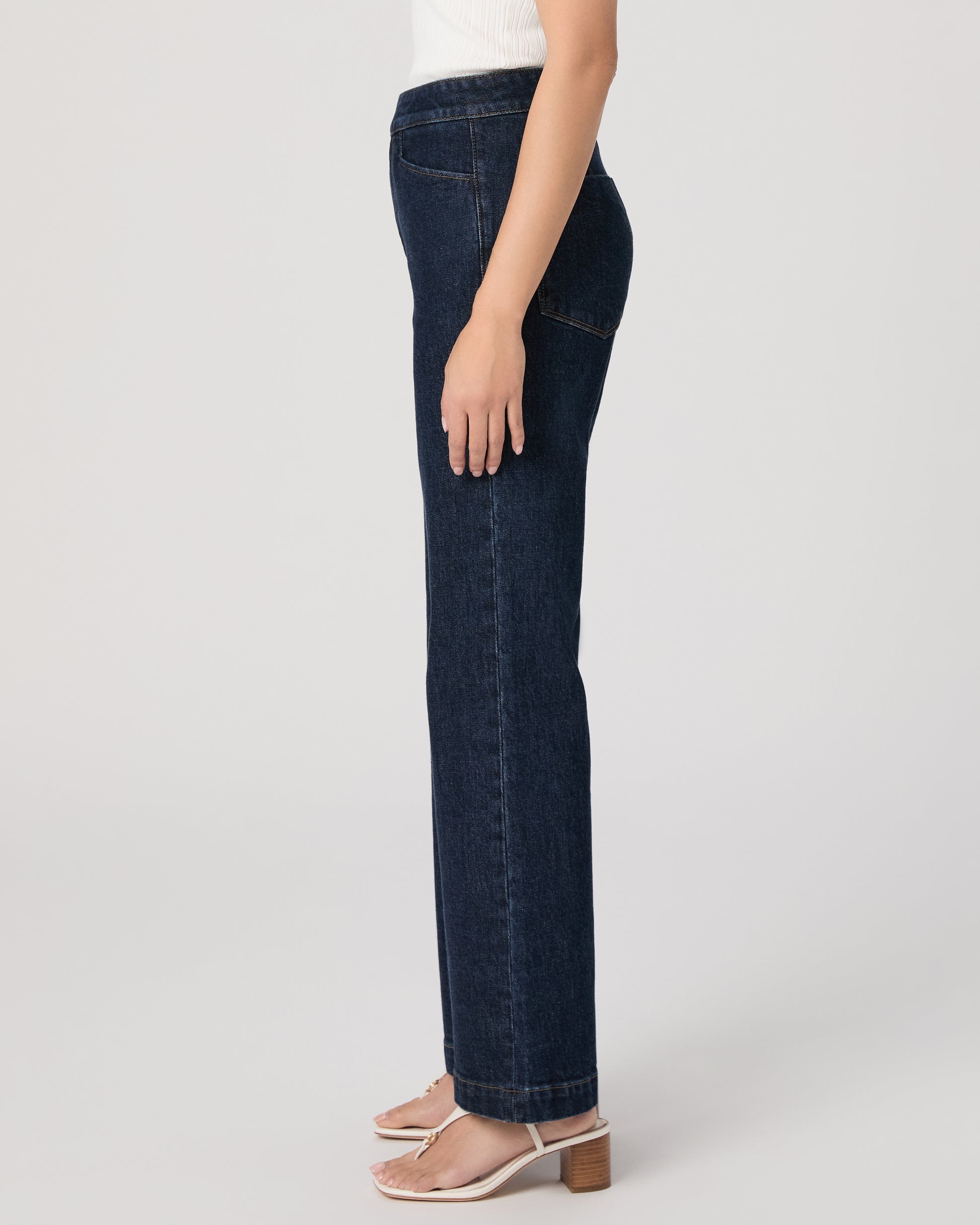 Woman wearing a white sleeveless top and dark blue jeans paired with white sandals on a white background