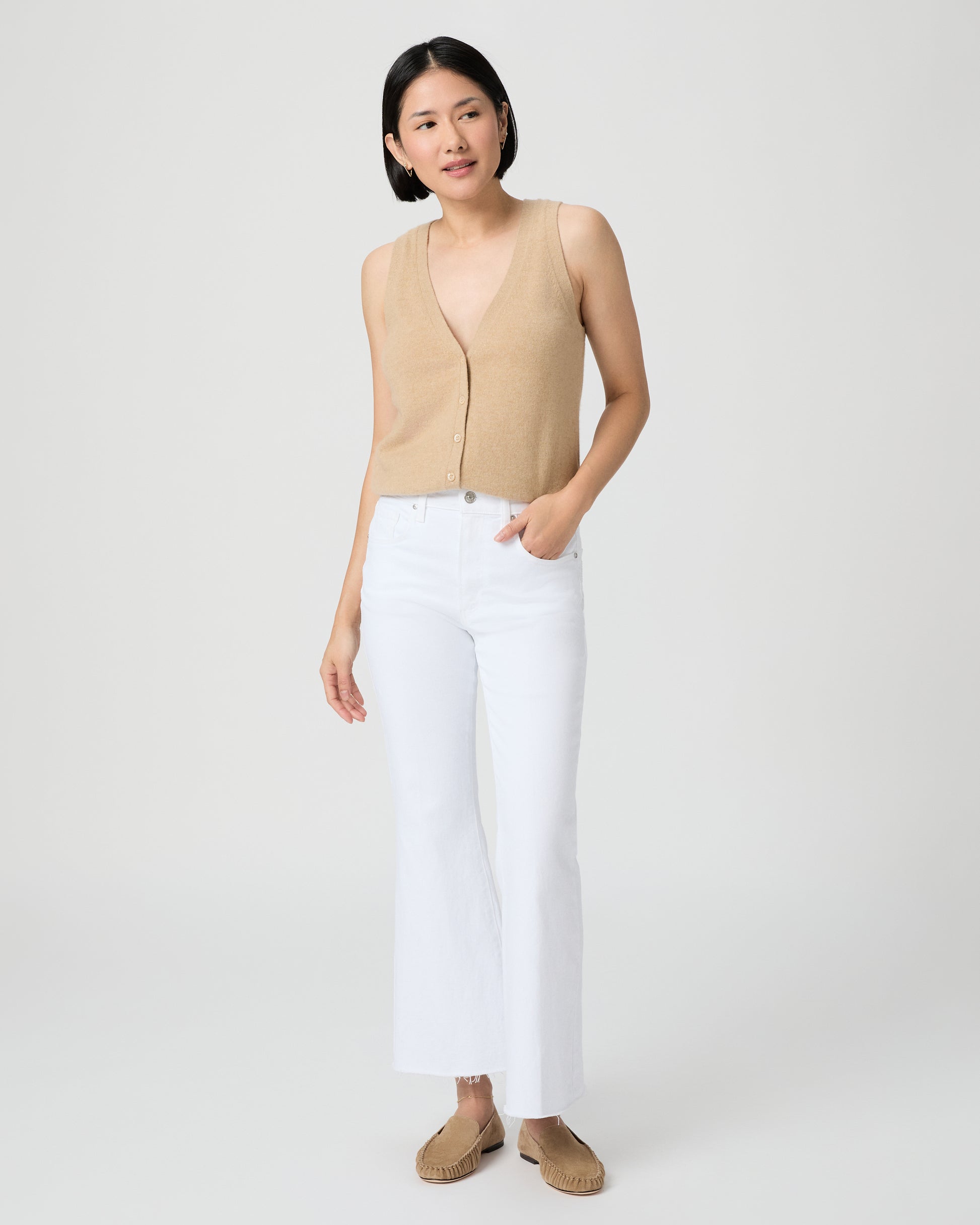 Woman wearing a beige sleeveless top and white pants on a plain background
