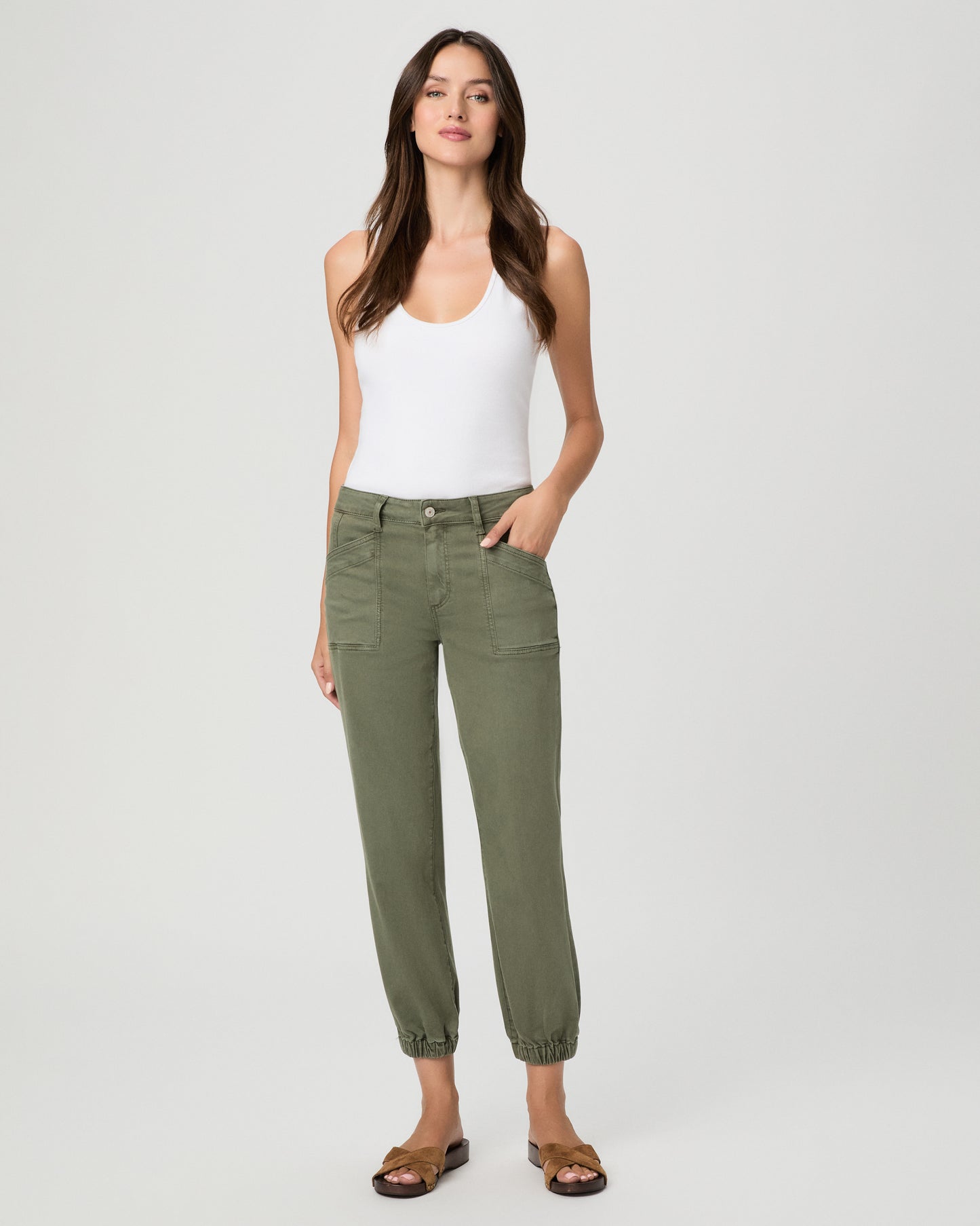 A woman modeling a pair of olive green cropped pants with a white tank top and brown sandals, standing against a light gray studio background.