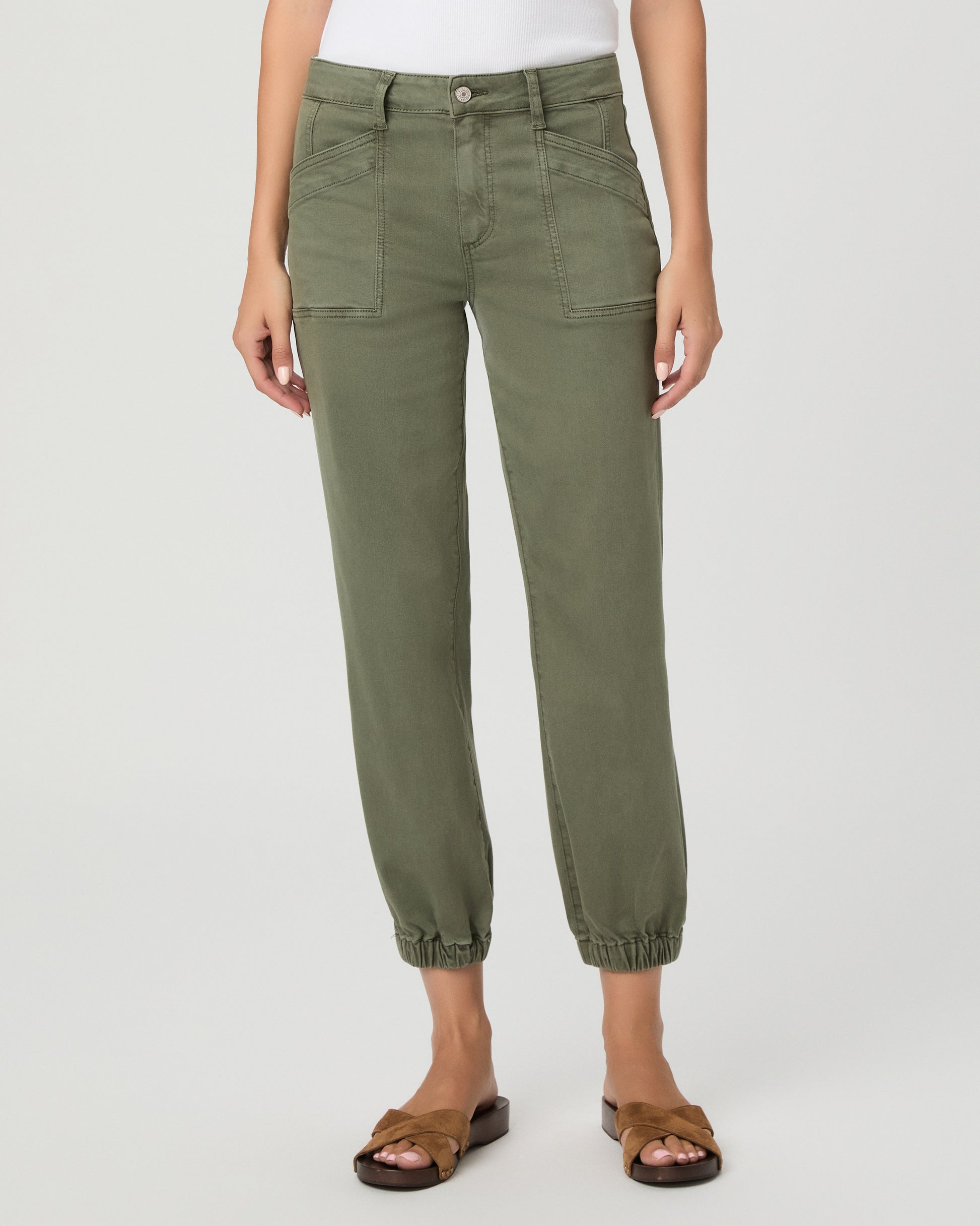 A woman modeling a pair of olive green cropped pants with a white tank top and brown sandals, standing against a light gray studio background.