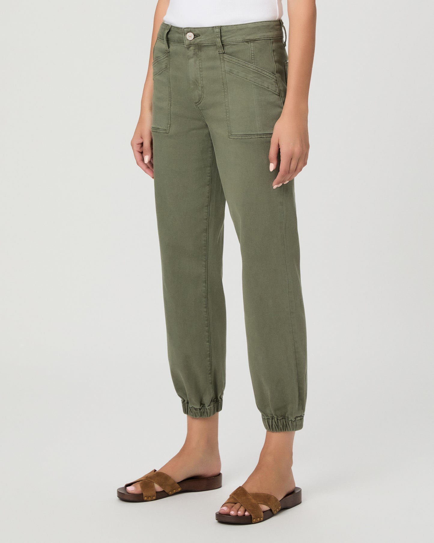 A woman modeling a pair of olive green cropped pants with a white tank top and brown sandals, standing against a light gray studio background.