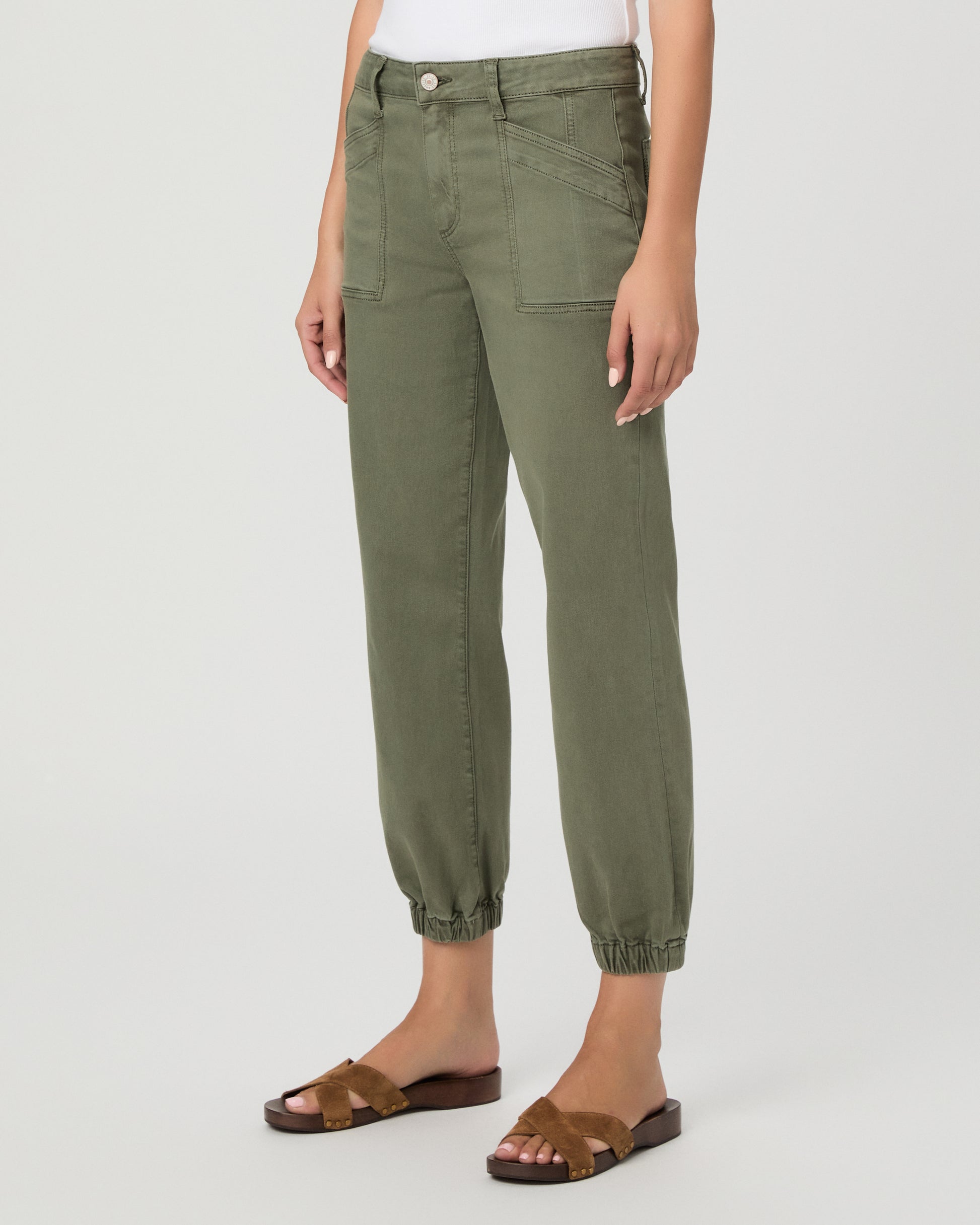 A woman modeling a pair of olive green cropped pants with a white tank top and brown sandals, standing against a light gray studio background.