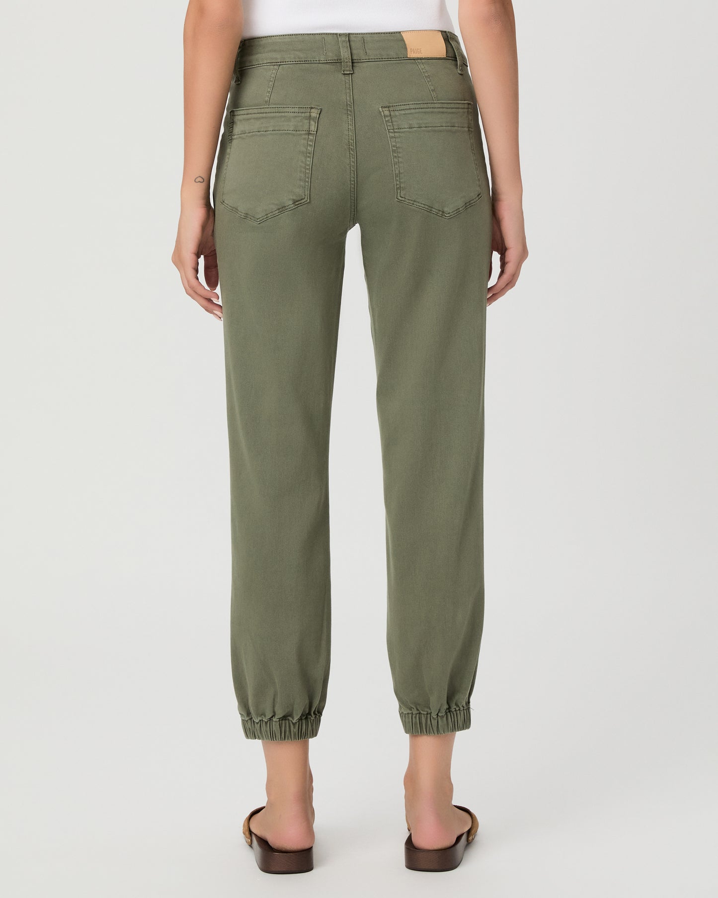 A woman modeling a pair of olive green cropped pants with a white tank top and brown sandals, standing against a light gray studio background.