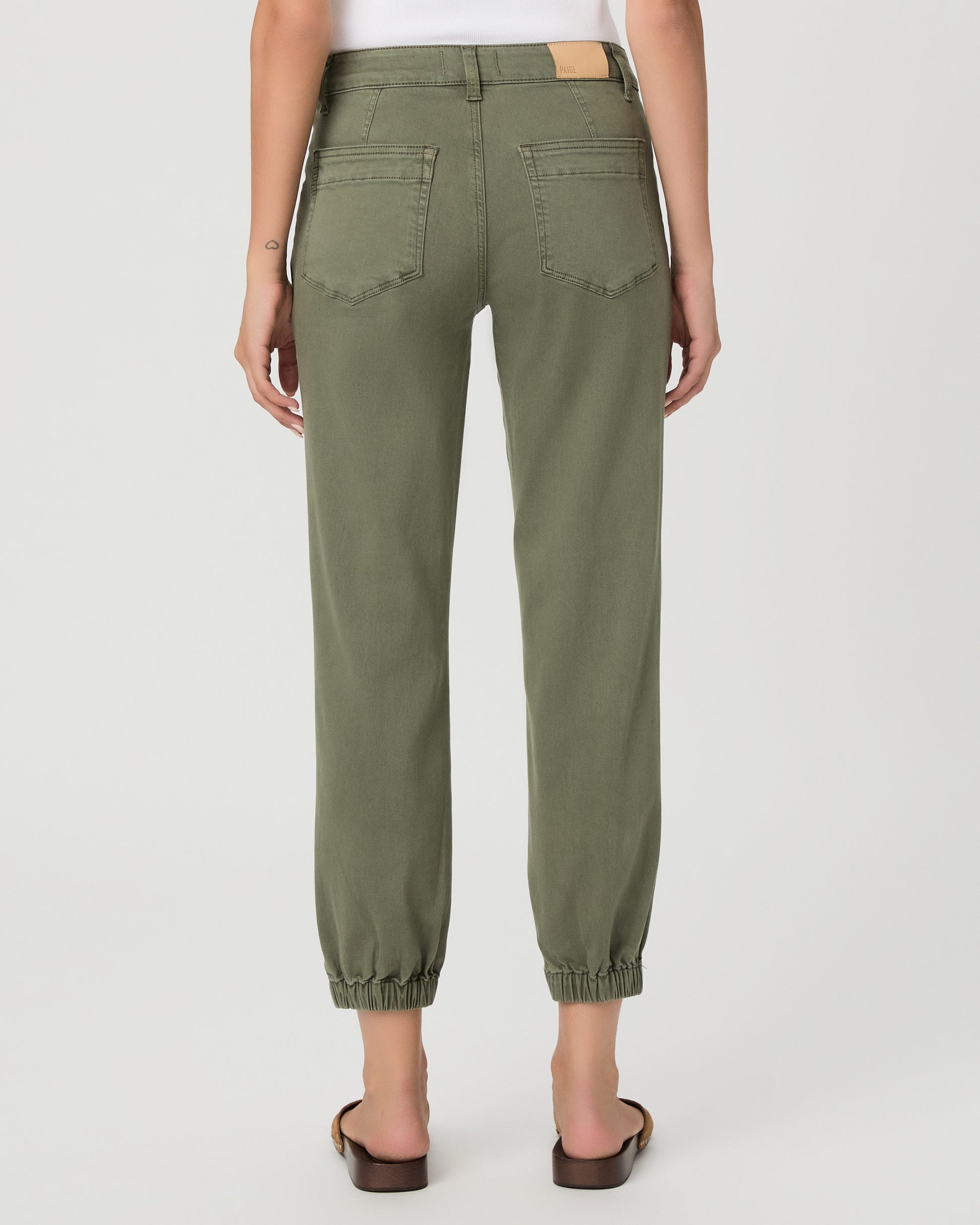 A woman modeling a pair of olive green cropped pants with a white tank top and brown sandals, standing against a light gray studio background.