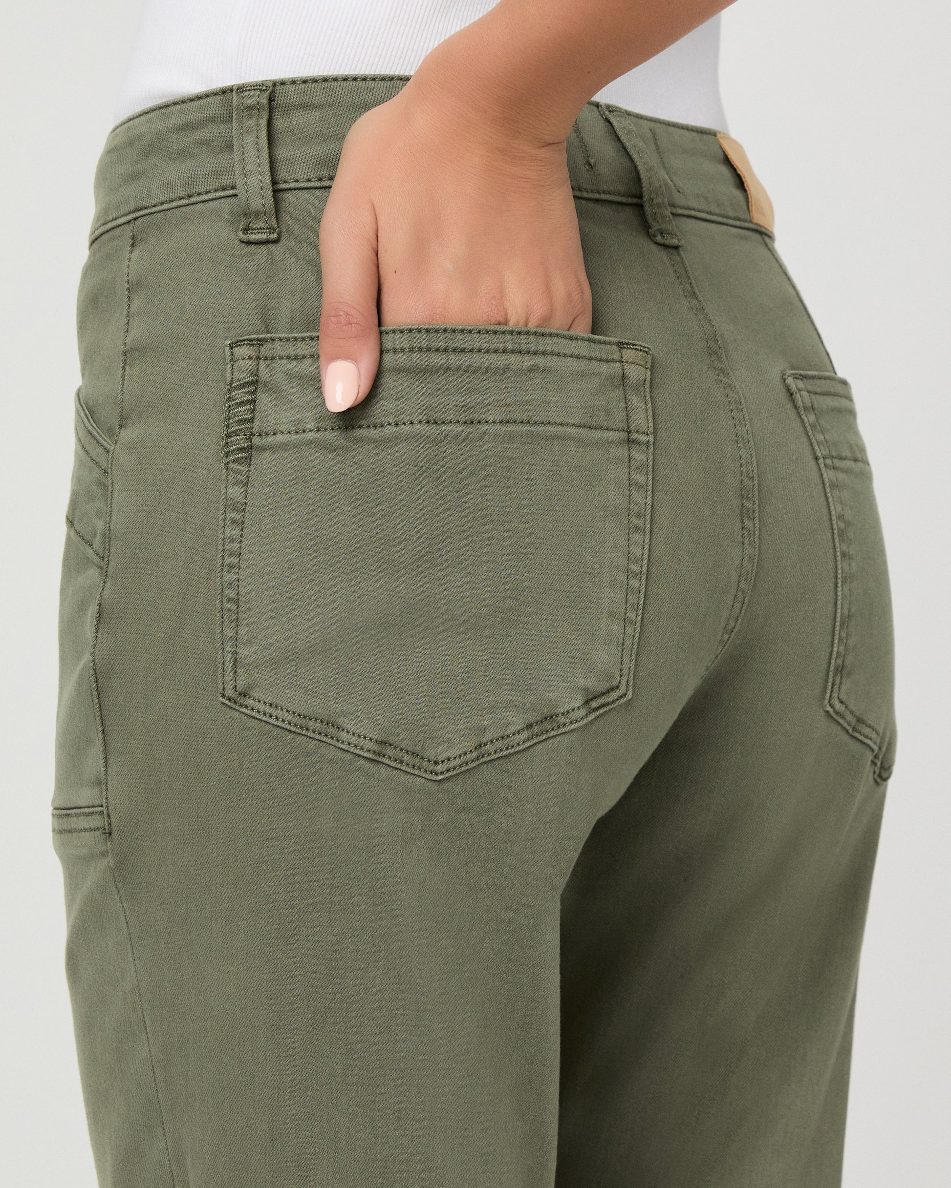 A woman modeling a pair of olive green cropped pants with a white tank top and brown sandals, standing against a light gray studio background.