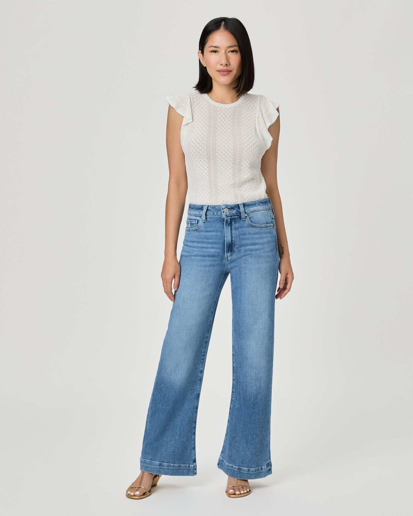 A woman modeling a white textured sleeveless top with ruffle details and high-waisted, wide-leg blue jeans against a light gray studio background.