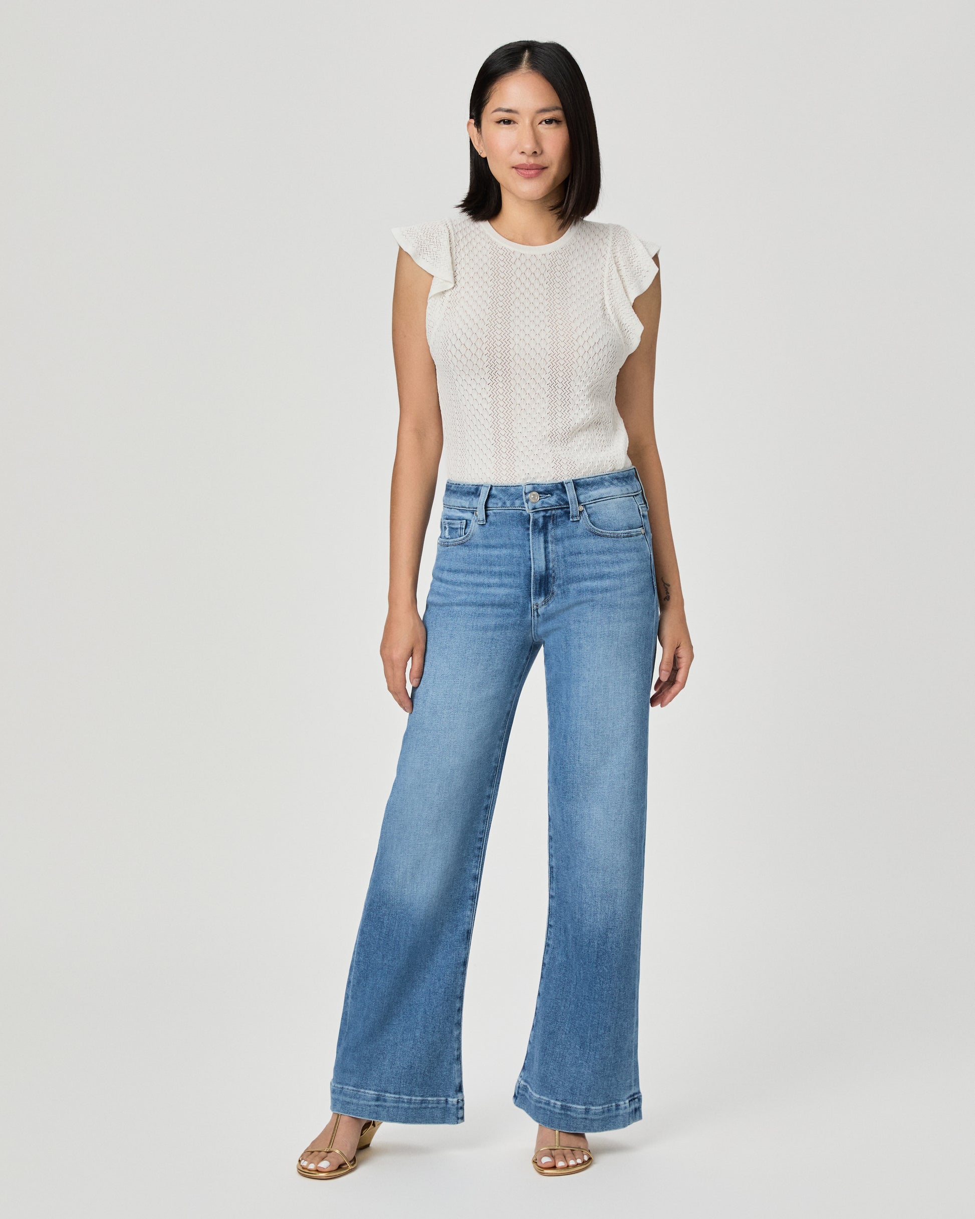 A woman modeling a white textured sleeveless top with ruffle details and high-waisted, wide-leg blue jeans against a light gray studio background.