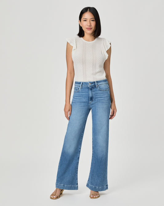 A woman modeling a white textured sleeveless top with ruffle details and high-waisted, wide-leg blue jeans against a light gray studio background.