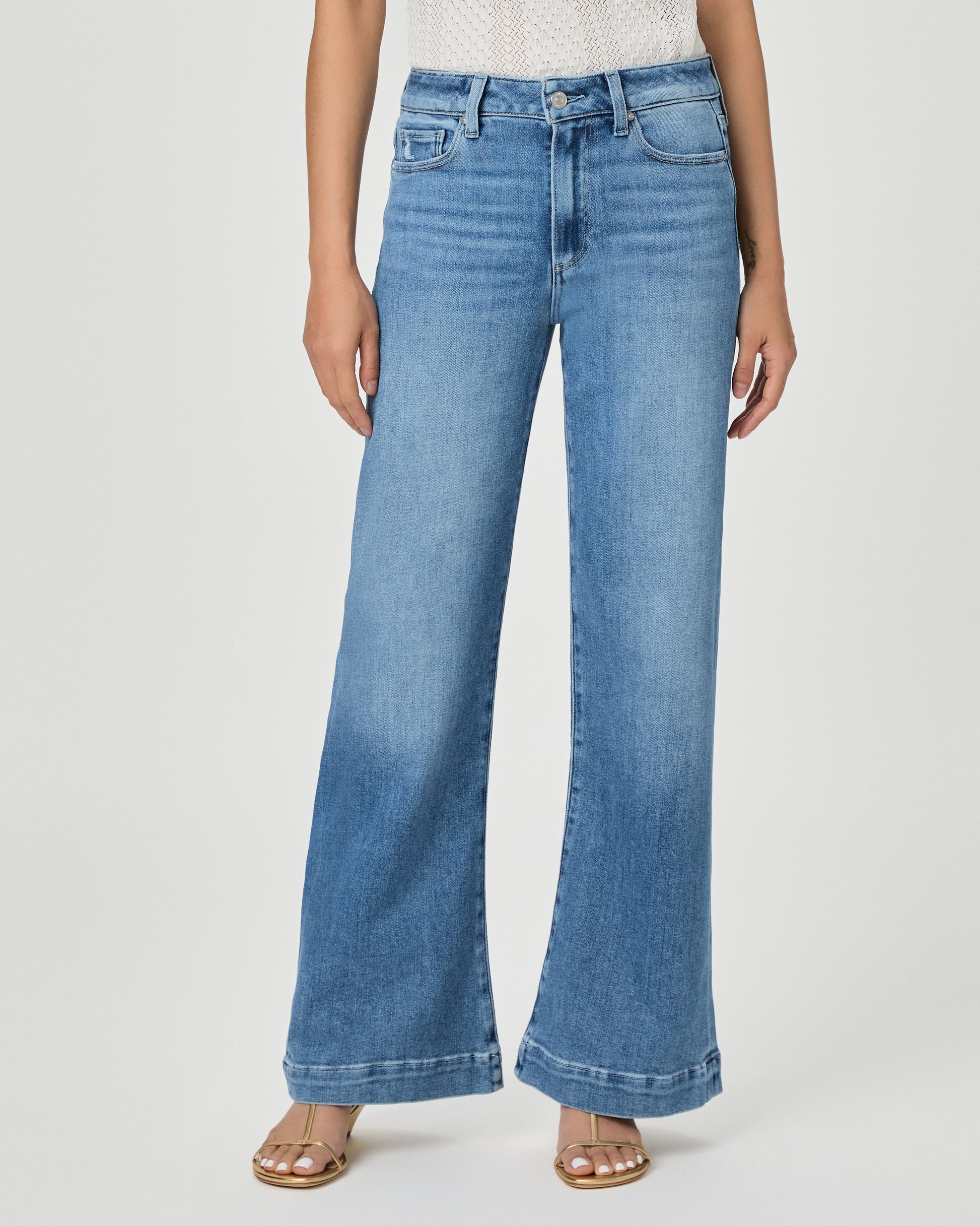 A woman modeling a white textured sleeveless top with ruffle details and high-waisted, wide-leg blue jeans against a light gray studio background.