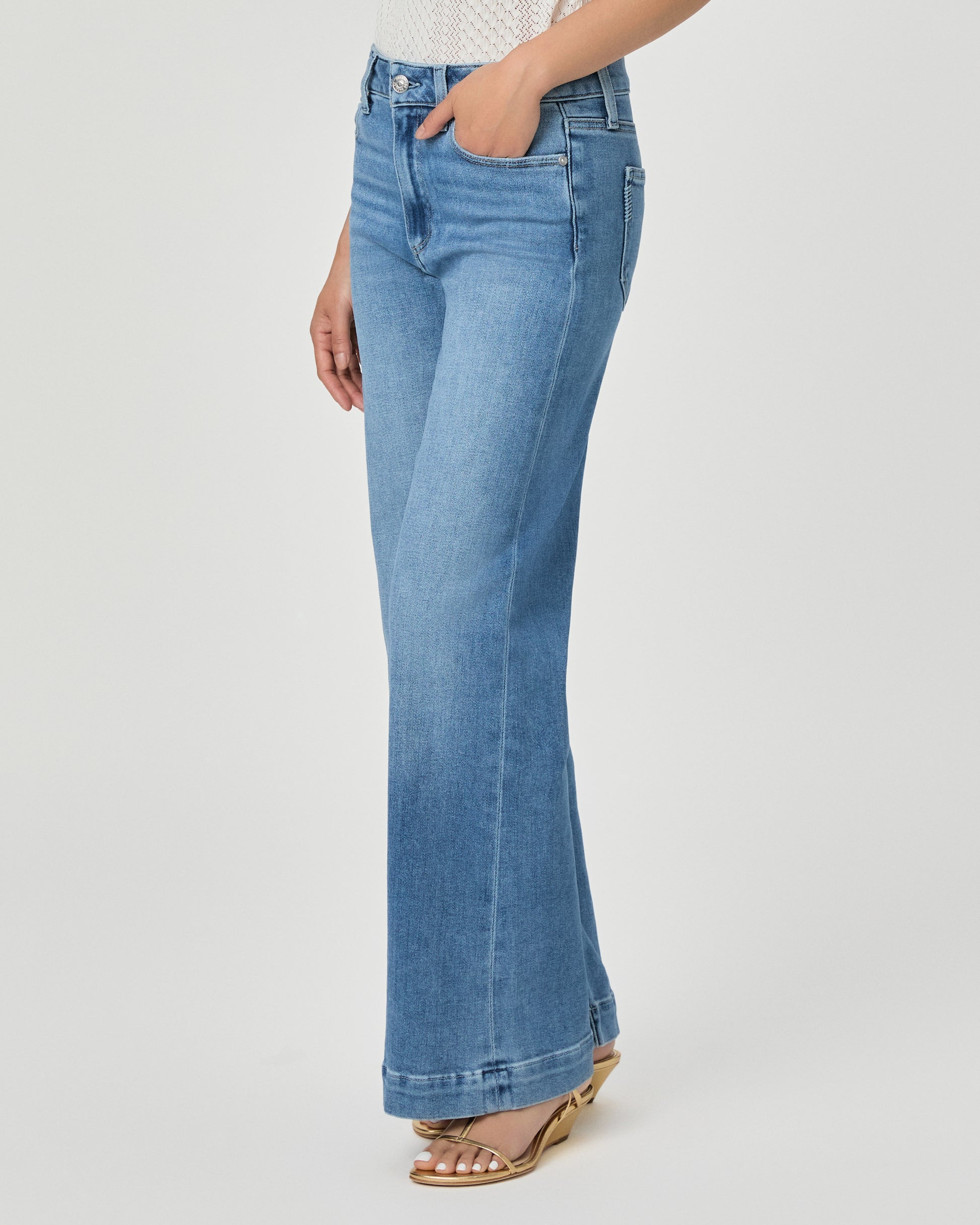 A woman modeling a white textured sleeveless top with ruffle details and high-waisted, wide-leg blue jeans against a light gray studio background.