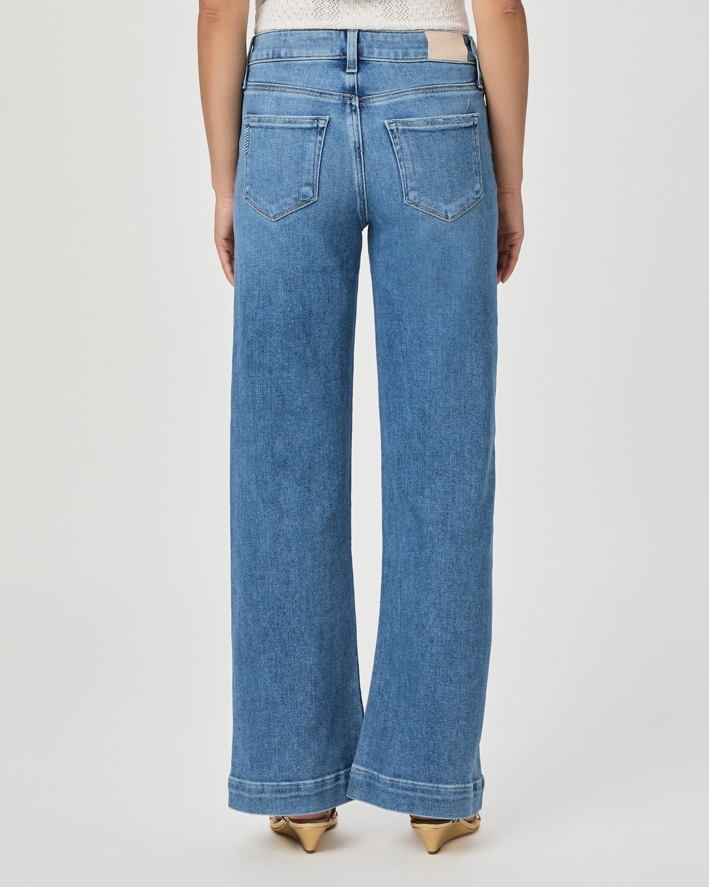 A woman modeling a white textured sleeveless top with ruffle details and high-waisted, wide-leg blue jeans against a light gray studio background.