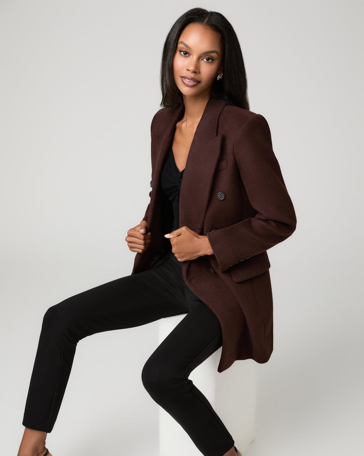 Close-up of a woman in a brown tailored blazer and black slim trousers, crouching with one knee bent and a hand near her collar.