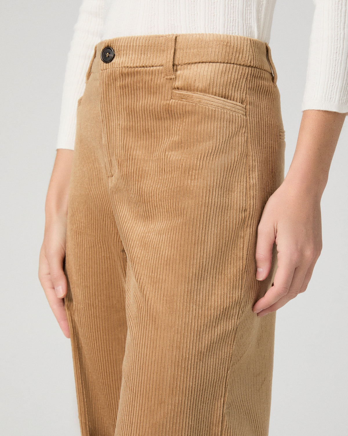 A woman modeling a white long-sleeve knit top with lace trim and tan wide-leg corduroy pants against a light gray studio background.