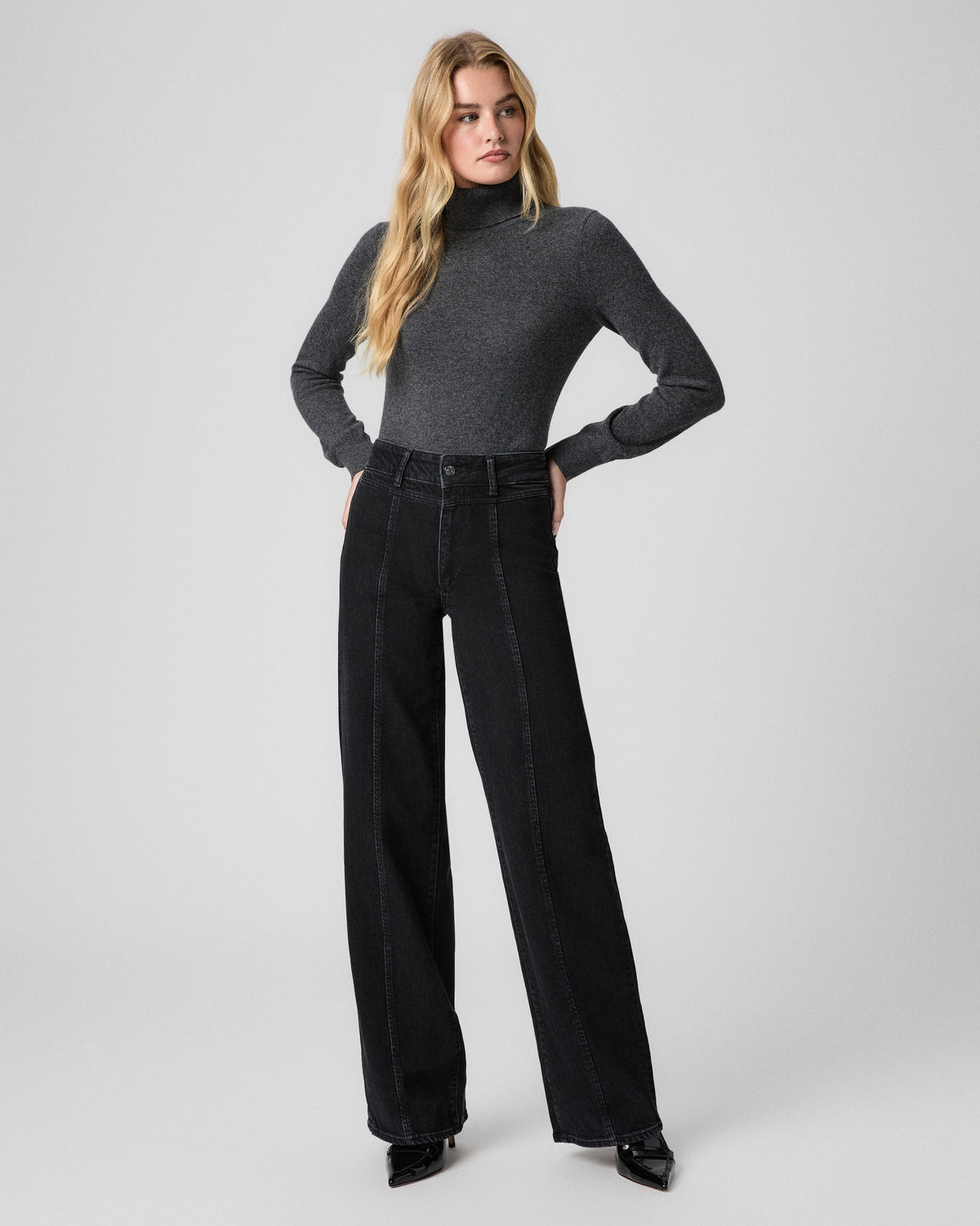Sasha Wide Leg Jean in Downtown Noir black with front seams; model stands facing forward with hands on hips, in a grey turtleneck and black pointed heels