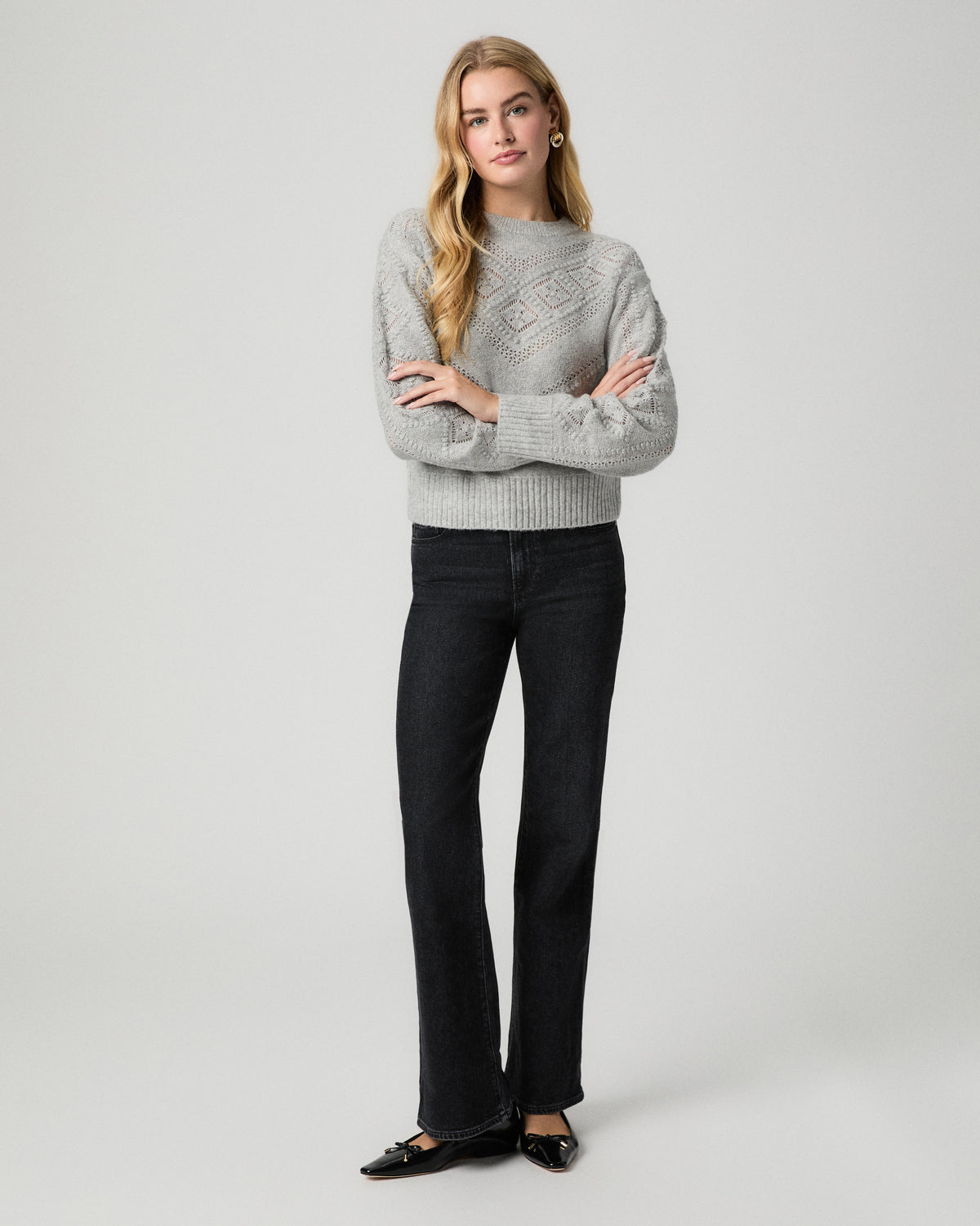 Full-length view of model standing with arms crossed, wearing the Sutter Sweater in Light Heather Grey with black straight jeans and black flats
