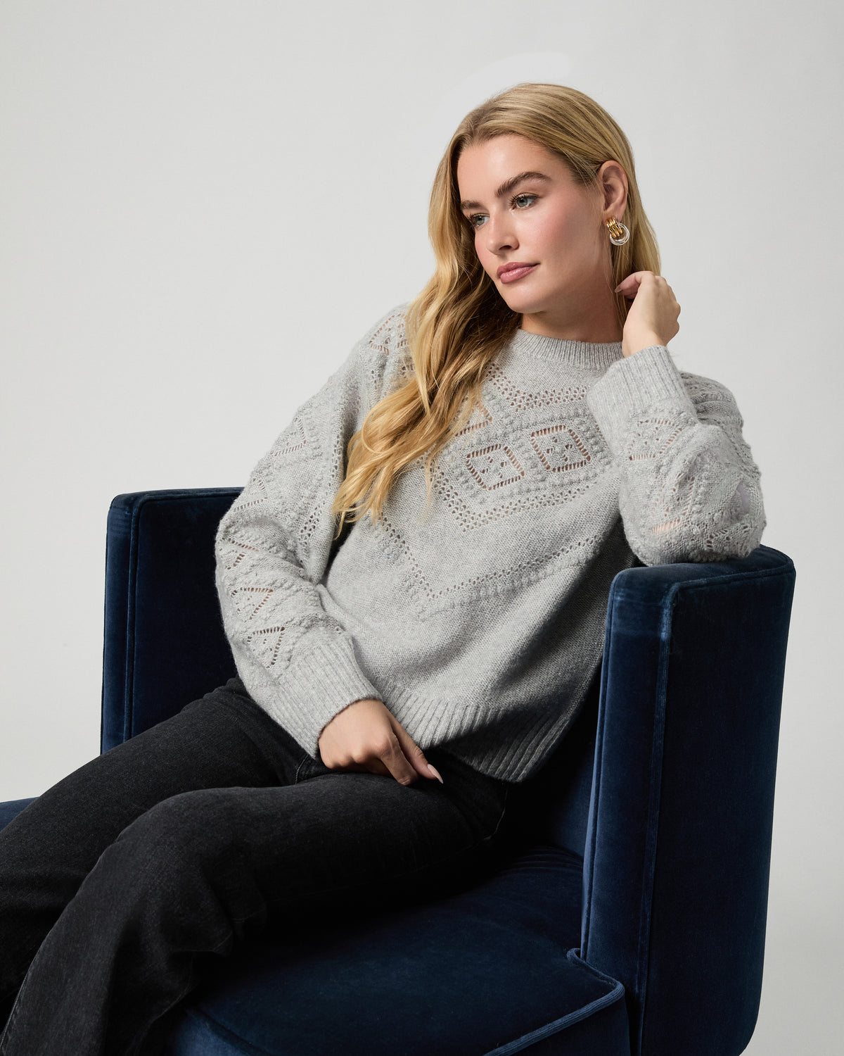 Model seated in a navy velvet armchair with one hand at her ear, wearing the Sutter Sweater in Light Heather Grey with black jeans