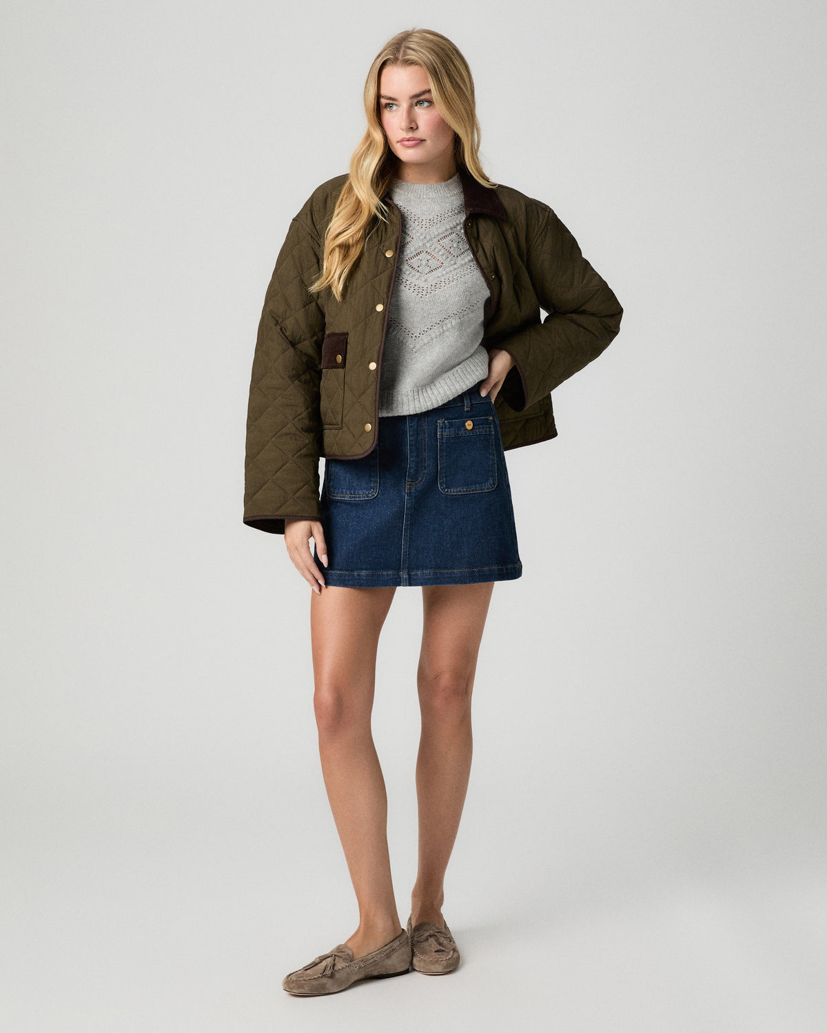 Full-length view of model with one hand in her pocket, wearing an olive jacket over the Sutter Sweater in Light Heather Grey with a dark denim mini skirt and tan flats