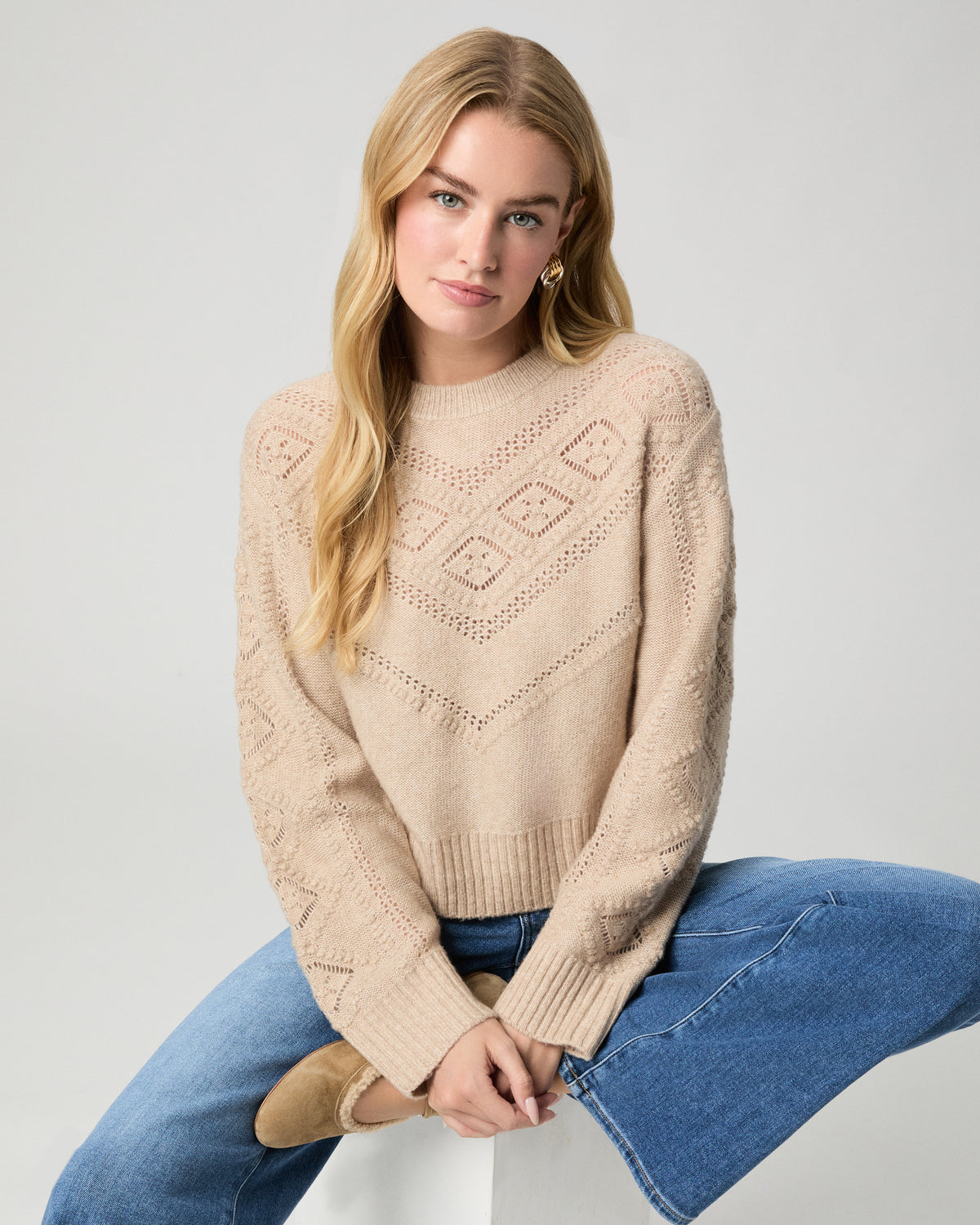 Model seated on a white block with knees raised, wearing the Sutter Sweater in Oatmeal Heather, a pointelle-knit crewneck, with medium-wash jeans and tan suede boots