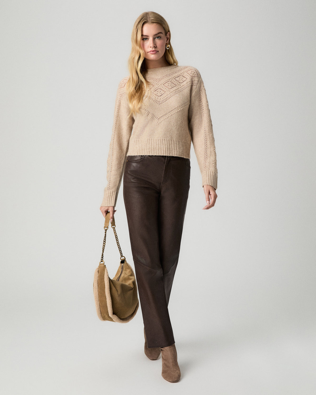 Full-length view of model walking with a tan bag, wearing the Sutter Sweater in Oatmeal Heather with dark brown coated straight jeans
