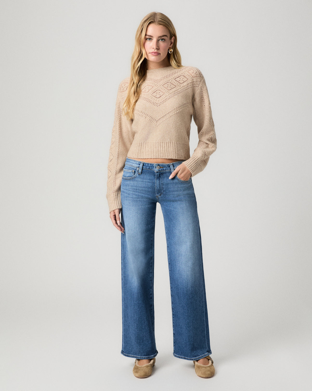 Full-length view of model with one hand in her pocket, wearing the Sutter Sweater in Oatmeal Heather with medium-wash wide-leg jeans and tan flats