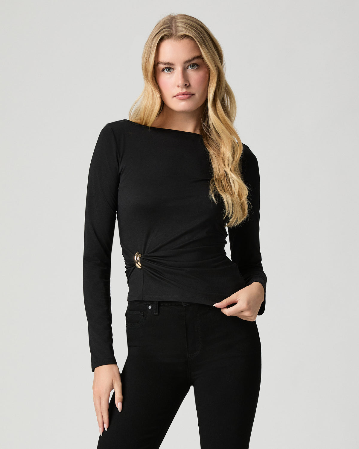 Model standing with one hand at her waist, wearing the Marianna Top in Black, a fitted long-sleeve boat neck with side hardware detail, with black jeans