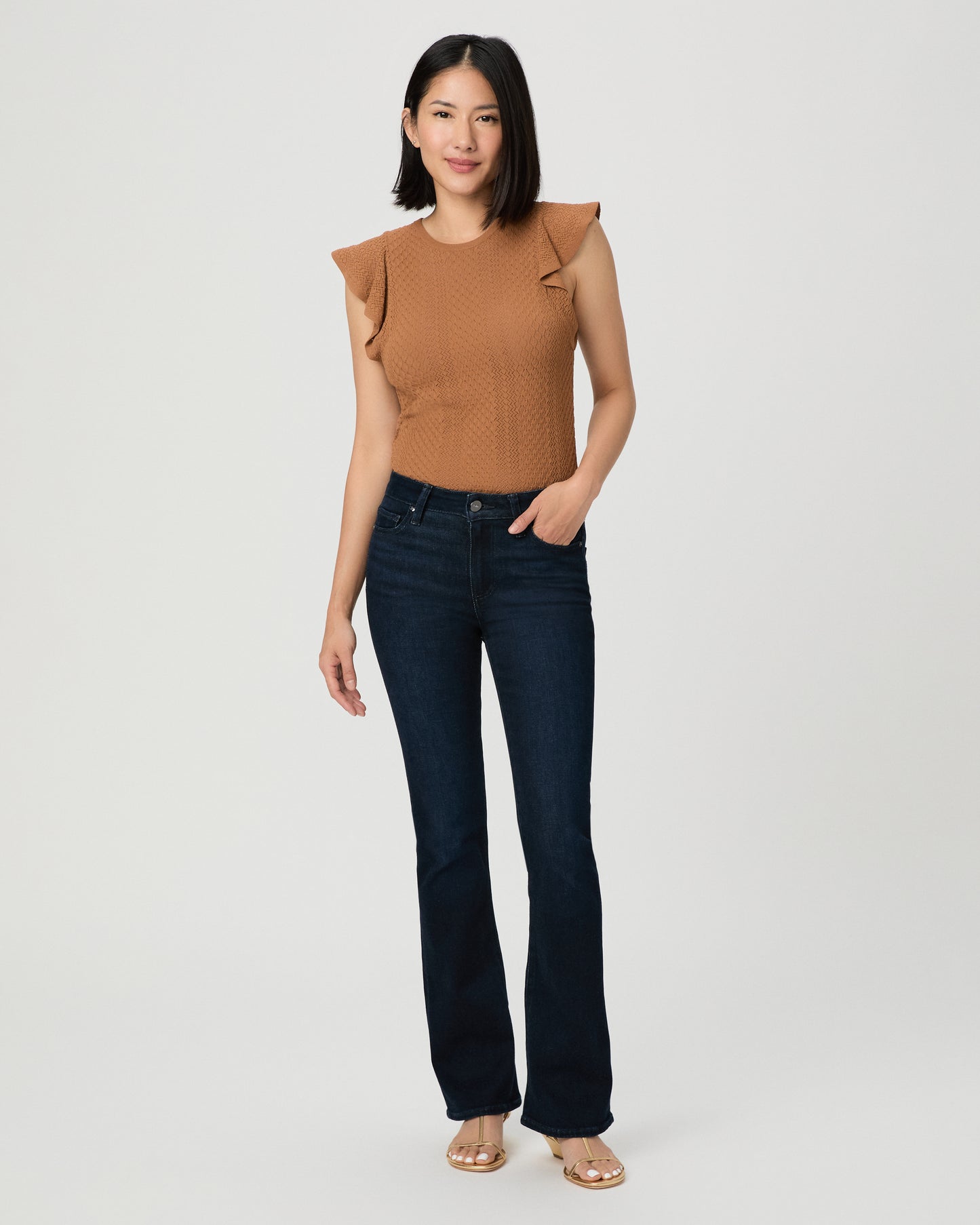 A woman modeling a sleeveless textured knit top in a warm camel brown with ruffled shoulder details, paired with dark blue jeans and beige sandals, against a light gray studio background.