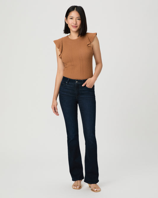 A woman modeling a sleeveless textured knit top in a warm camel brown with ruffled shoulder details, paired with dark blue jeans and beige sandals, against a light gray studio background.