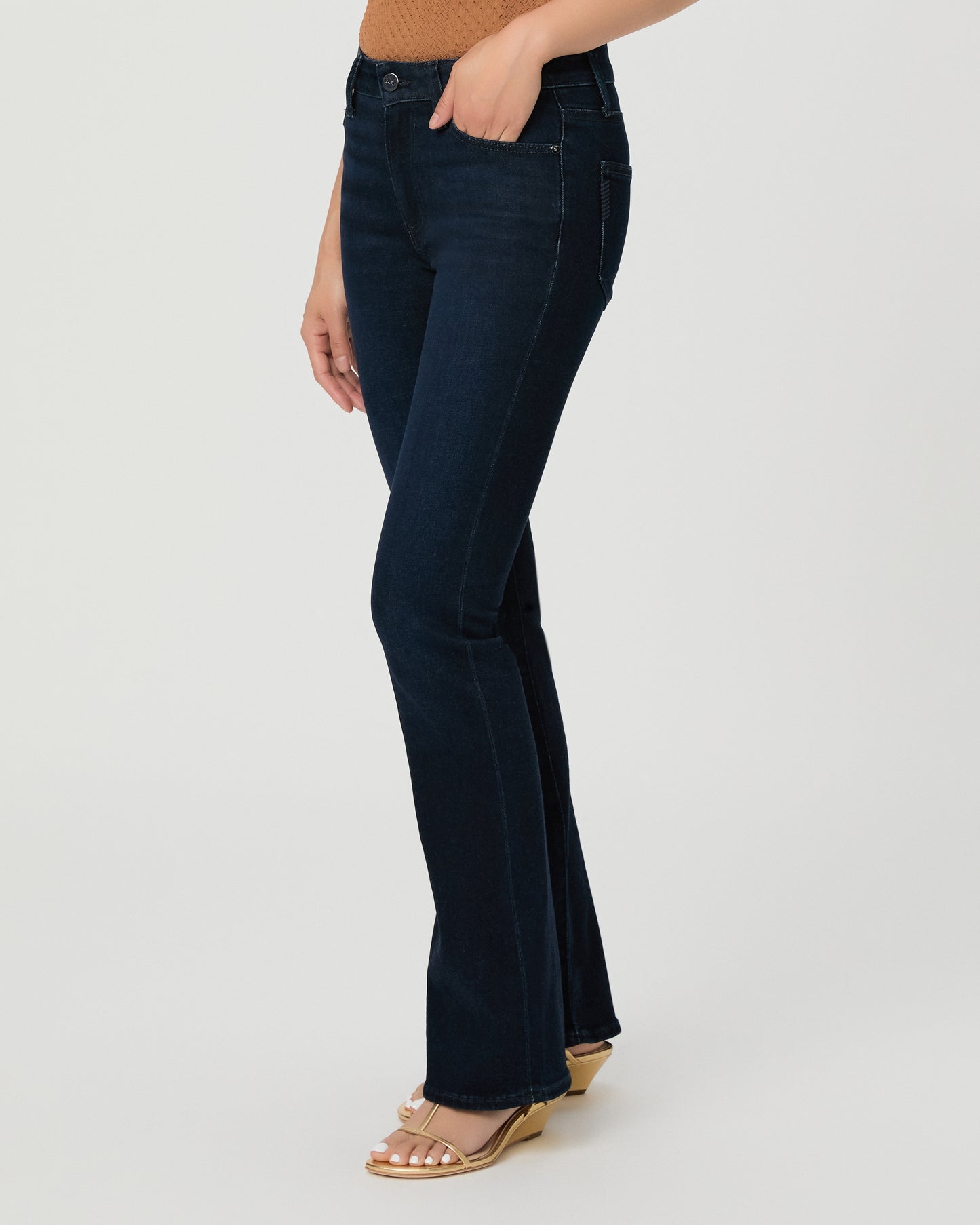 A woman modeling a sleeveless textured knit top in a warm camel brown with ruffled shoulder details, paired with dark blue jeans and beige sandals, against a light gray studio background.