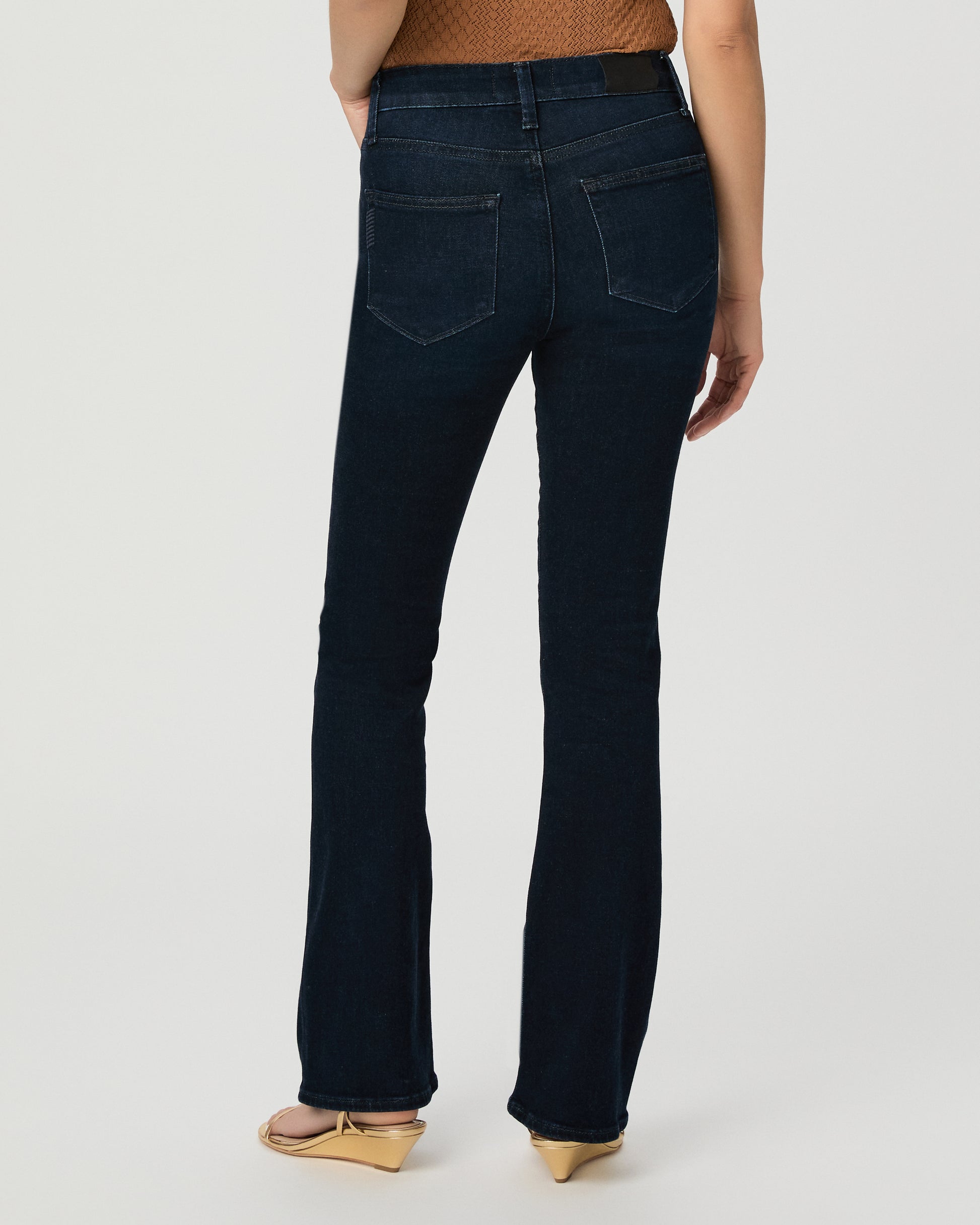 A woman modeling a sleeveless textured knit top in a warm camel brown with ruffled shoulder details, paired with dark blue jeans and beige sandals, against a light gray studio background.