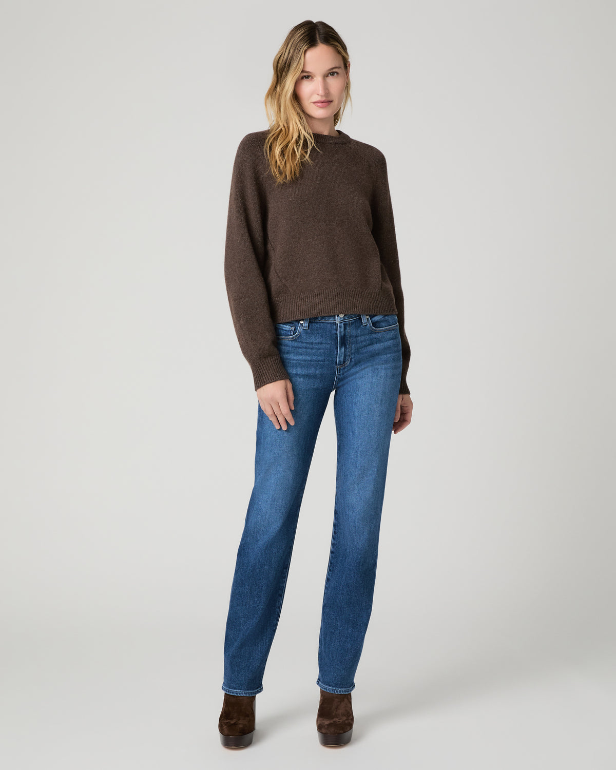 Women wearing a long sleeve brown sweater and blue jeans paired with brown boots against a white background