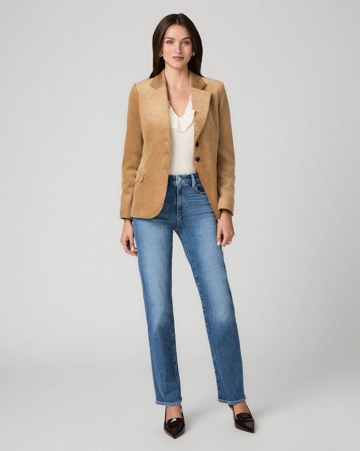 A woman modeling a tan suede blazer over a cream top and blue jeans, standing against a light gray studio background.