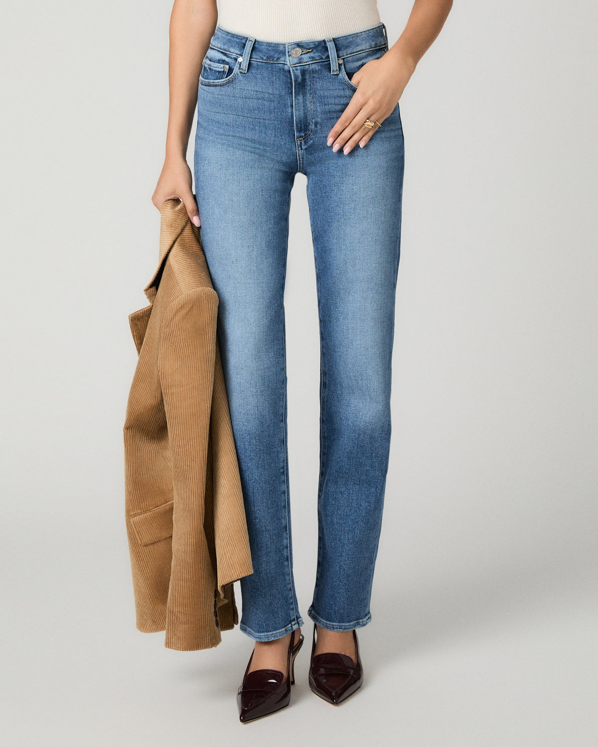 A model wearing medium-wash blue straight-leg jeans, styled with a tan blazer draped over one leg and dark brown pointed-toe flats, against a light gray studio background.