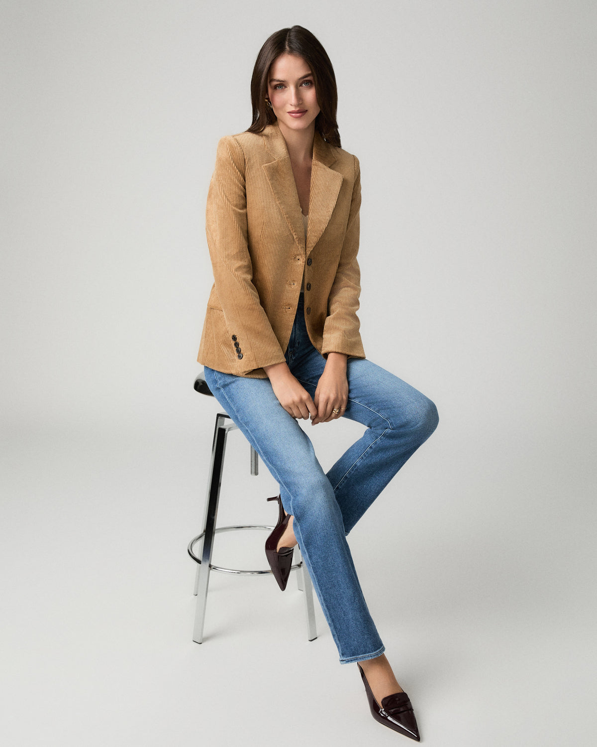 A woman sits on a metal stool wearing a tan corduroy blazer, blue jeans, and dark brown pointed-toe flats against a light gray studio background.