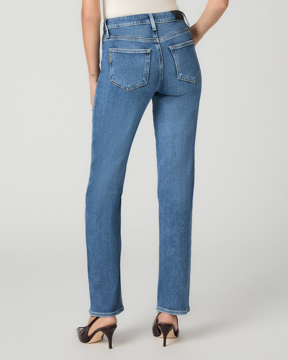 Rear view of a model wearing blue denim jeans with a straight-leg fit, paired with dark brown slingback high heels, against a light gray studio background.