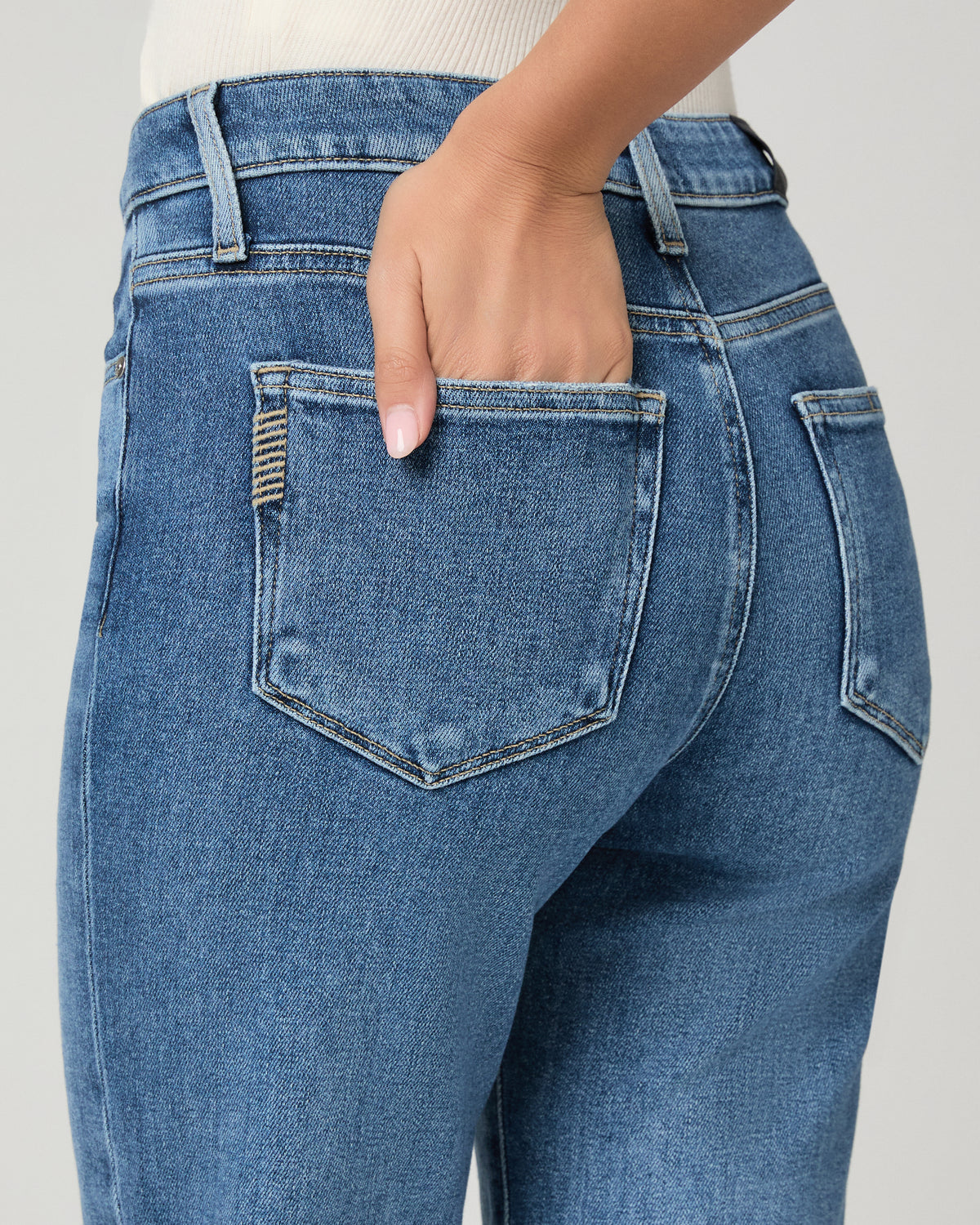 Close-up back view of a person wearing medium-wash blue denim jeans, with one hand tucked into the back pocket against a light gray studio background.
