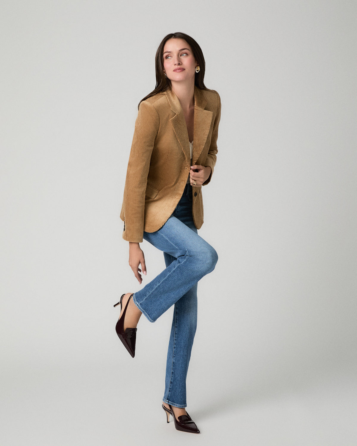 A woman posing in a studio wearing a tan suede blazer, blue jeans, and dark brown pointed-toe heels against a light gray background.