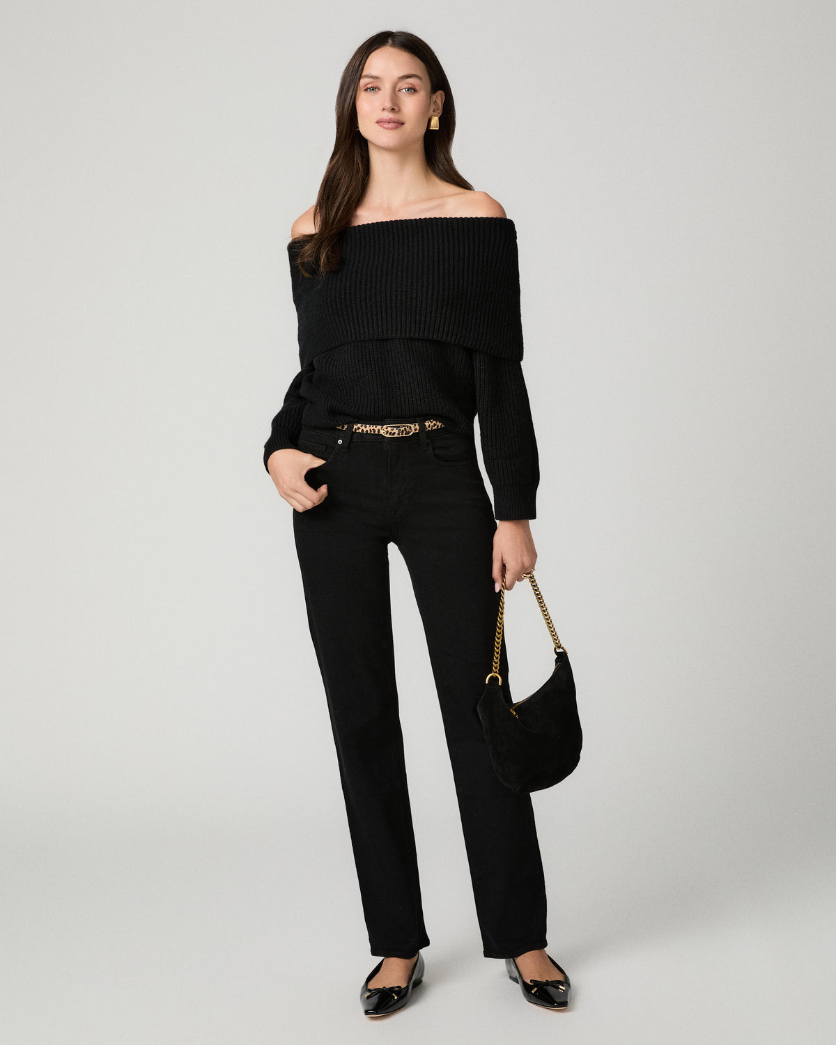 A woman models a black off-the-shoulder long-sleeve top paired with black straight-leg jeans, a gold chain belt, black pointed flats, and a black handbag with a gold chain strap, against a light gray studio background.
