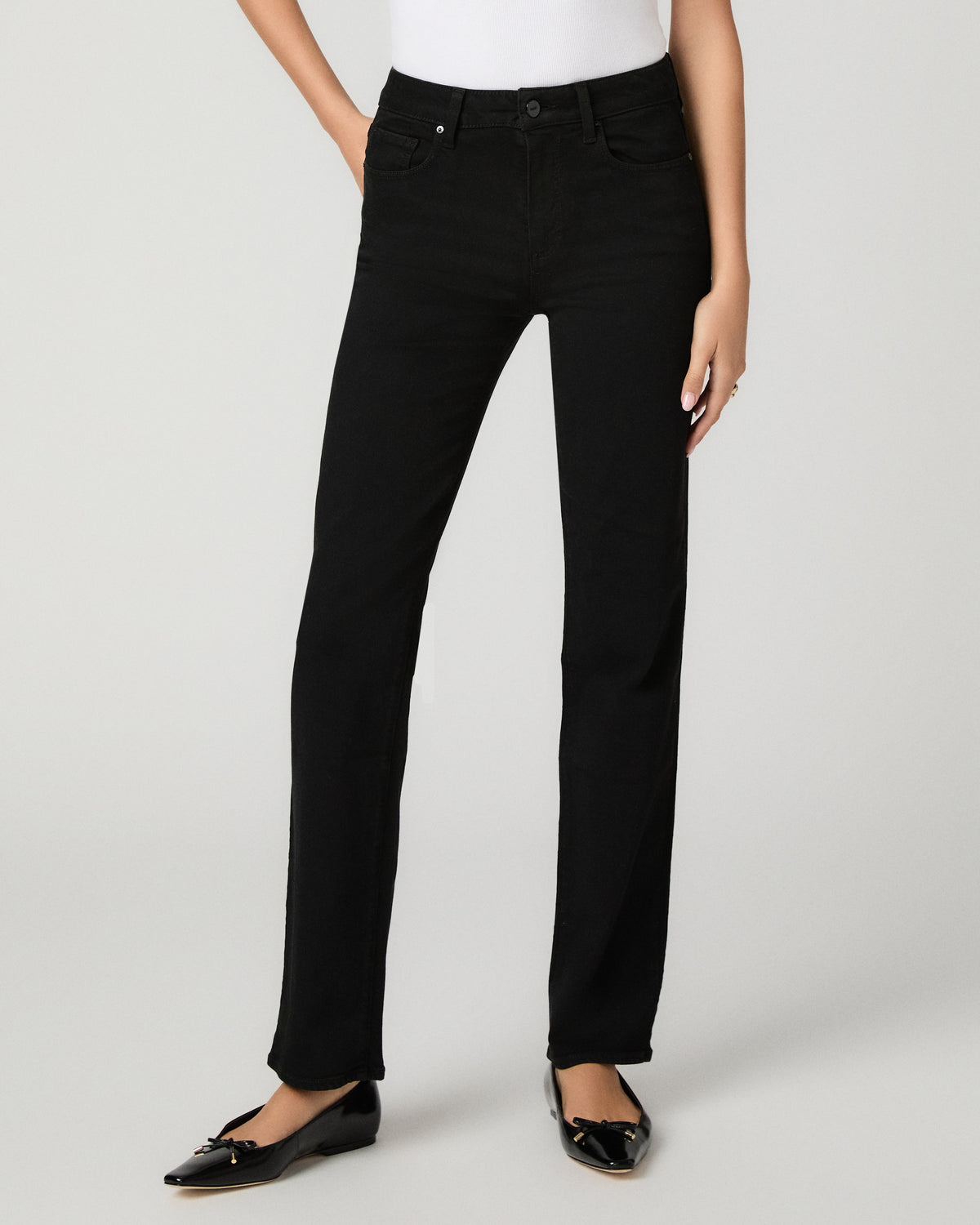 A model wearing black straight-leg jeans, styled with a white top and black patent leather flats, photographed against a light gray studio background.