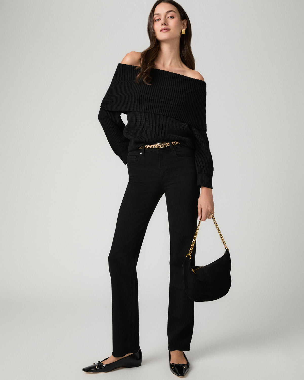 A model wearing a black off-the-shoulder ribbed knit top, black straight-leg jeans, and black flats, holding a black handbag with a gold chain strap against a light gray studio background.