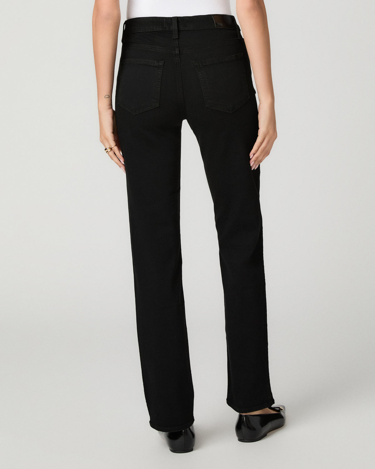 Rear view of a model wearing black straight-leg jeans, a white top, and black flats against a light gray studio background.