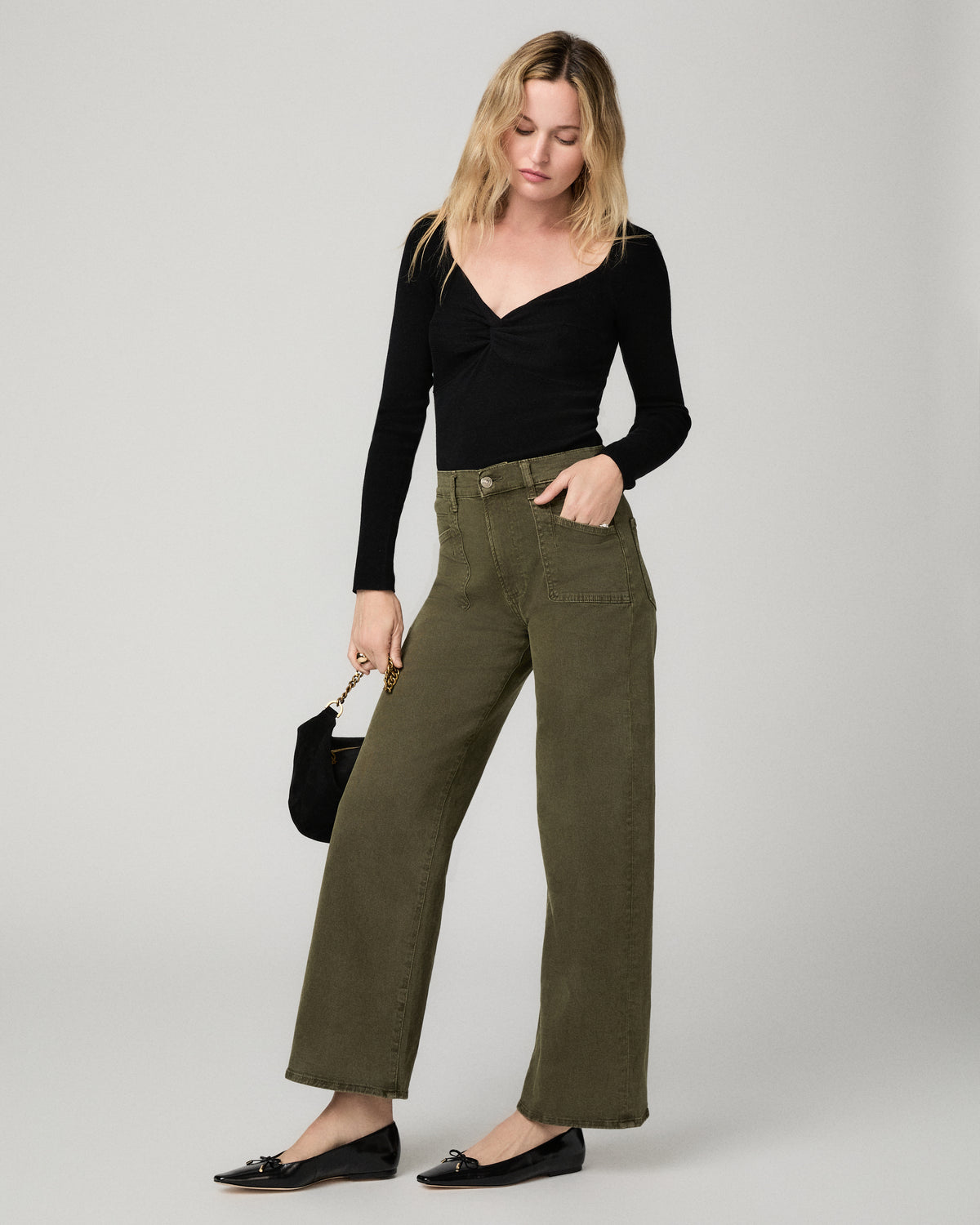 Full-length front view of a woman wearing the olive green Arellia Skimmer Barrel Leg Jean with a black V-neck sweater, black flats, and a black bag.