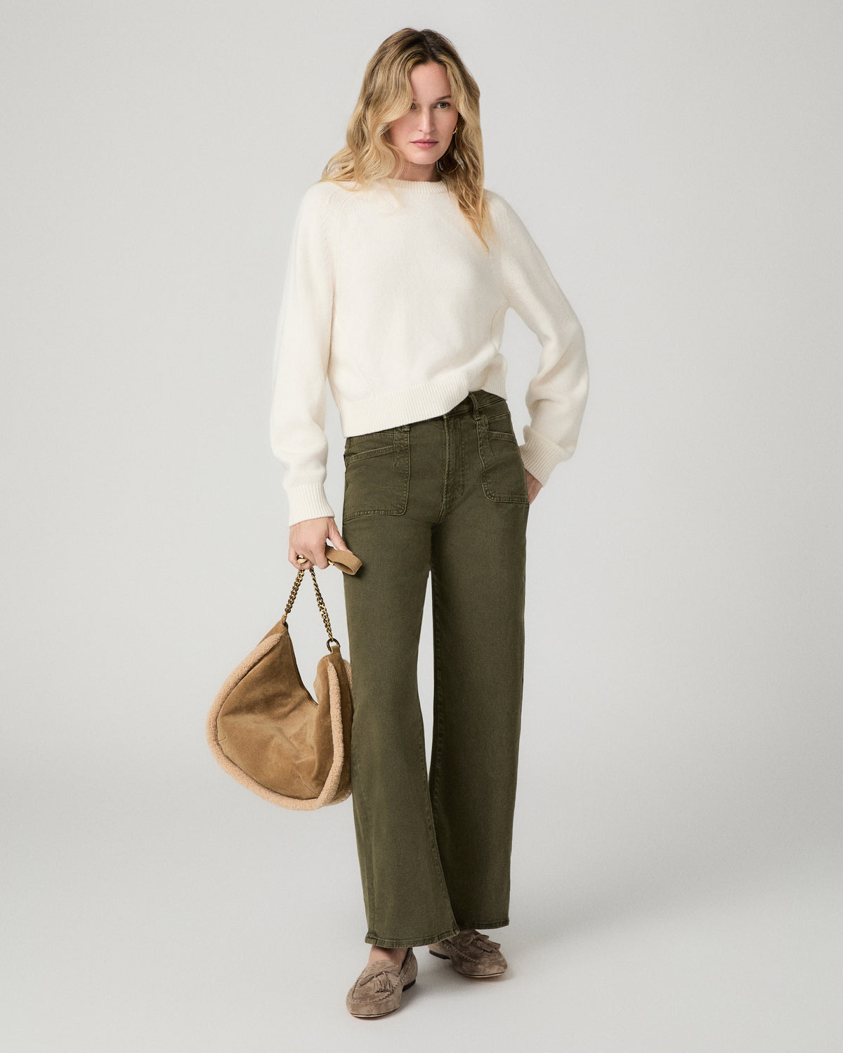 Full-length front view of a woman wearing the olive green Arellia Skimmer Barrel Leg Jean with a cream sweater, holding a tan bag.