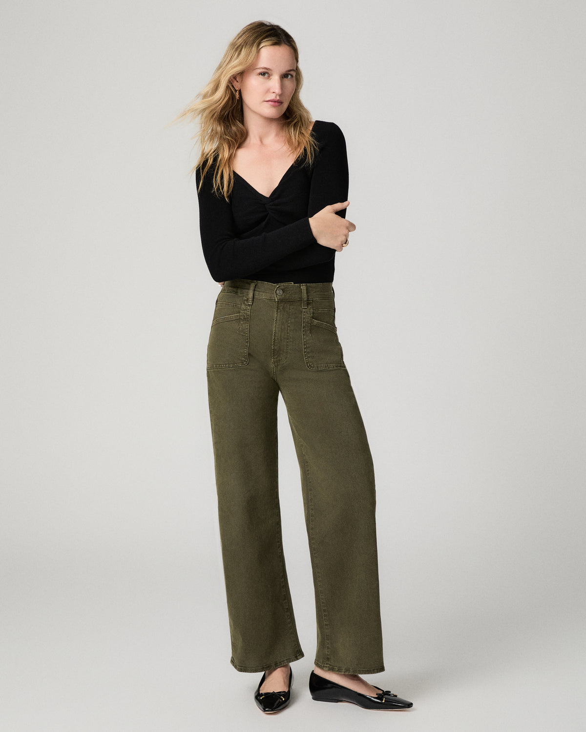Full-length front view of a woman wearing the olive green Arellia Skimmer Barrel Leg Jean with a black sweater, hand near her chin.