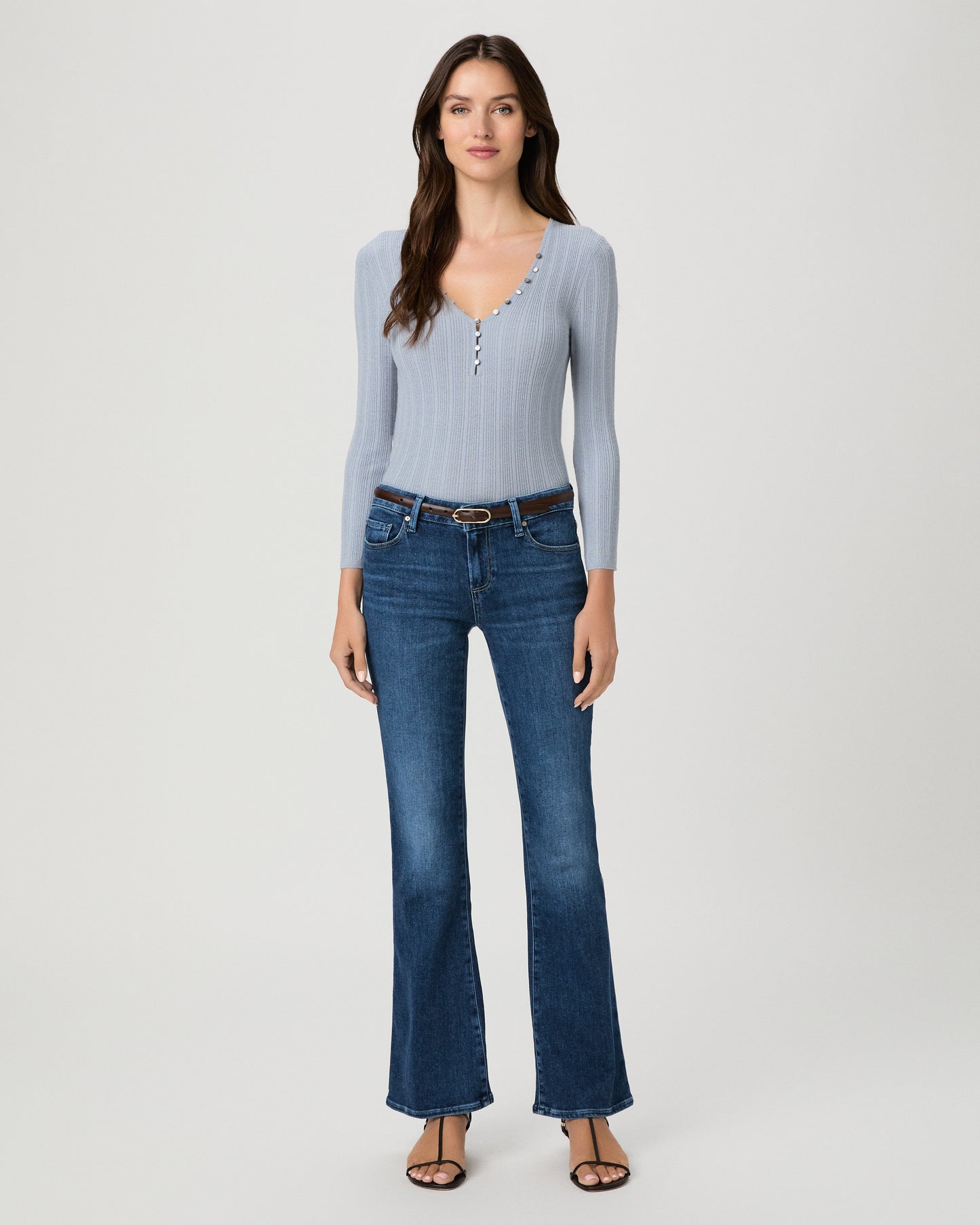 A woman modeling a light blue ribbed long-sleeve knit top with a buttoned henley neckline and dark blue flare jeans, standing against a light gray studio background.