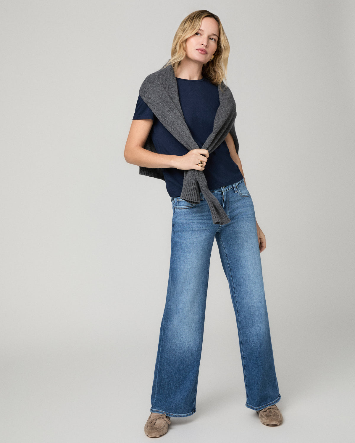 Women wearing a blue tee, grey sweater over shoulders paired with blue jeans standing against a white background