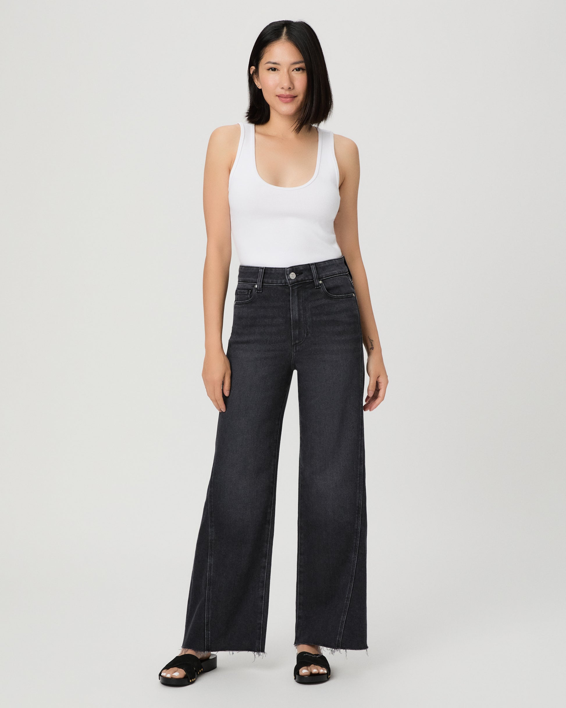 A woman modeling a pair of dark charcoal wide-leg jeans with a frayed hem, paired with a white tank top and black slide sandals, against a light gray studio background.